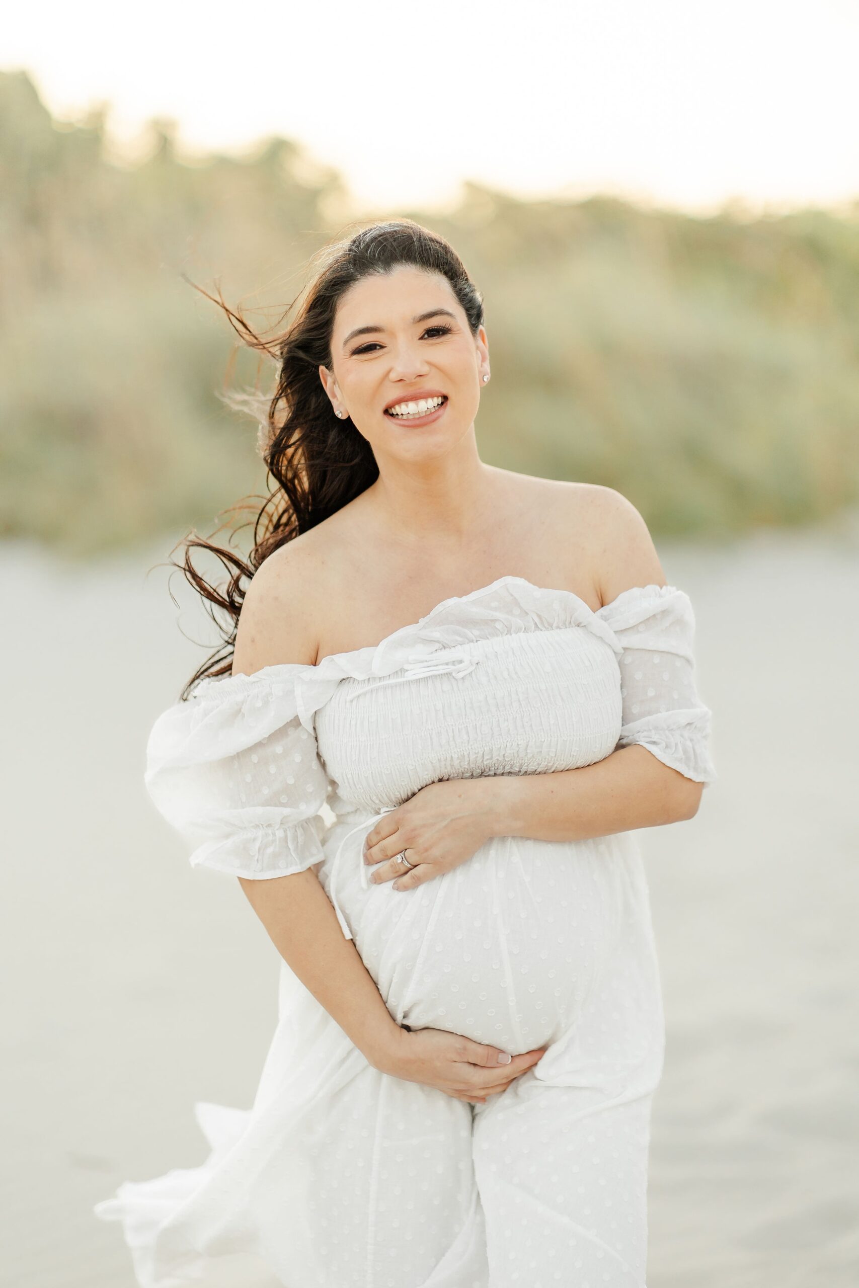 A smiling mother to be in a white maternity gown on a windy beach at sunset