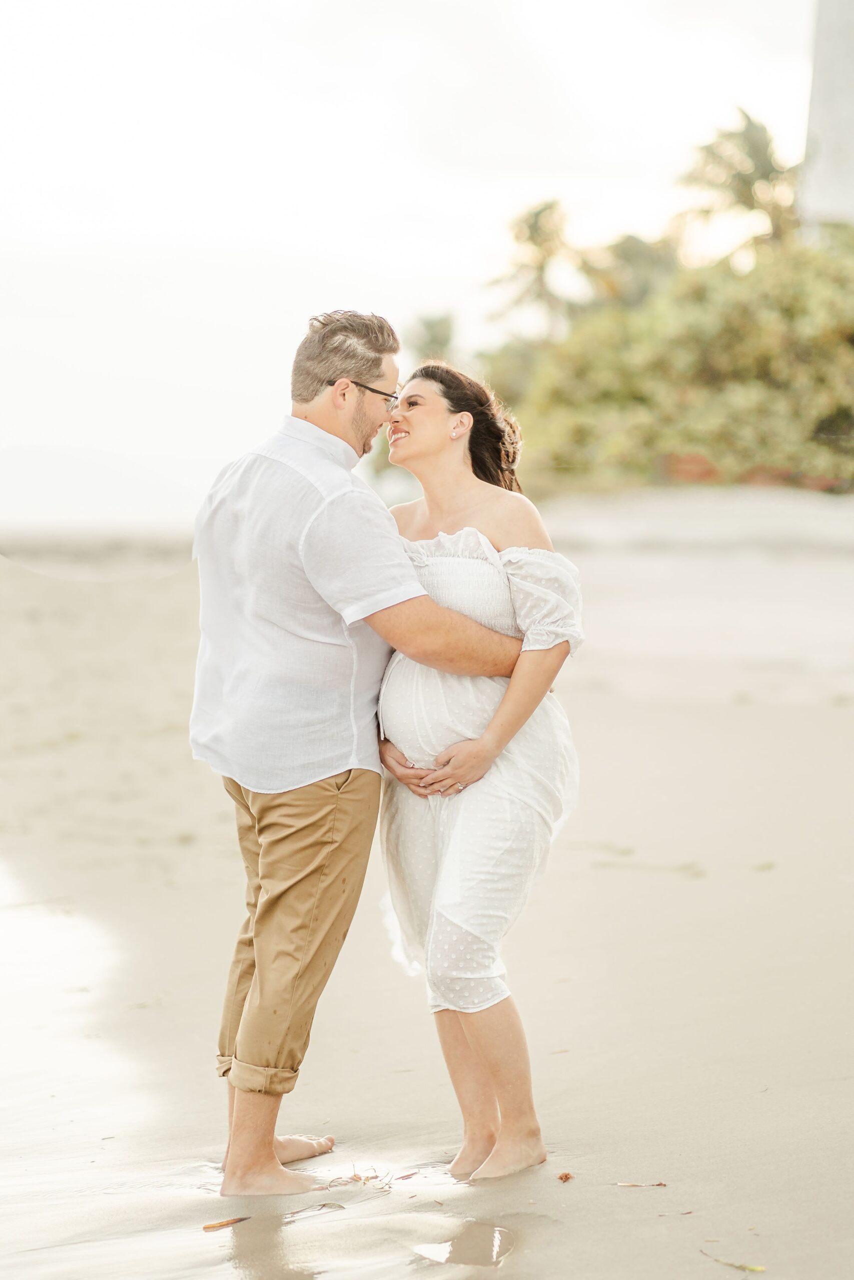 A pregnant couple leans in for a kiss while hugging on a windy beach at sunset after visiting south florida women's care