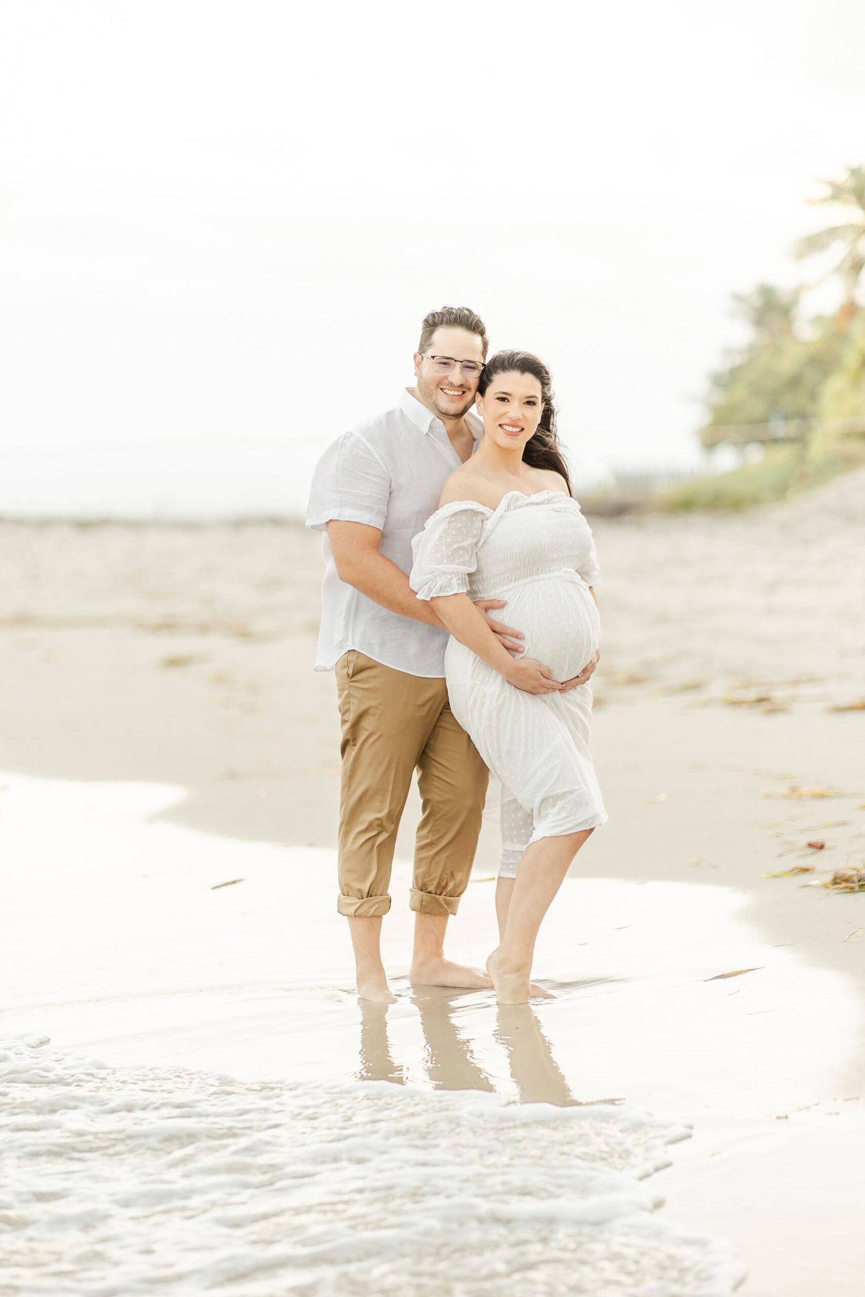 A happy couple snuggle and rest their hands on the bump while standing on a windy beach at sunset in white after visiting south florida women's care