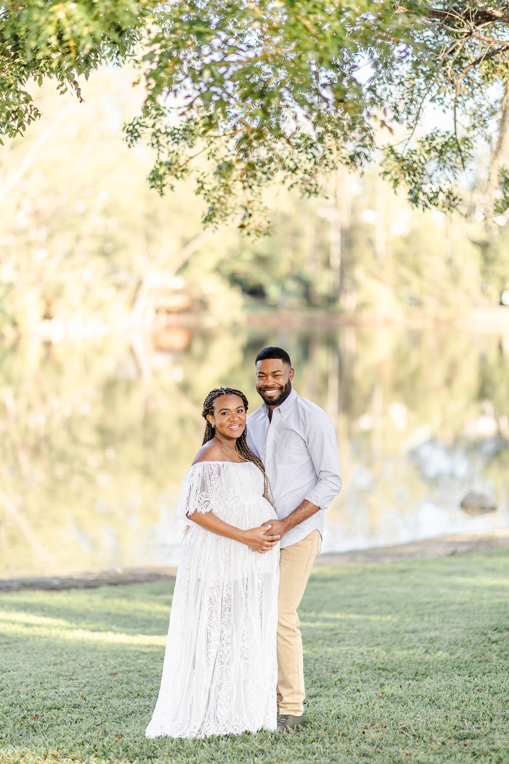 A happy expecting couple hold the bump while standing under a tree by a lake in a park after visiting tlc women's health