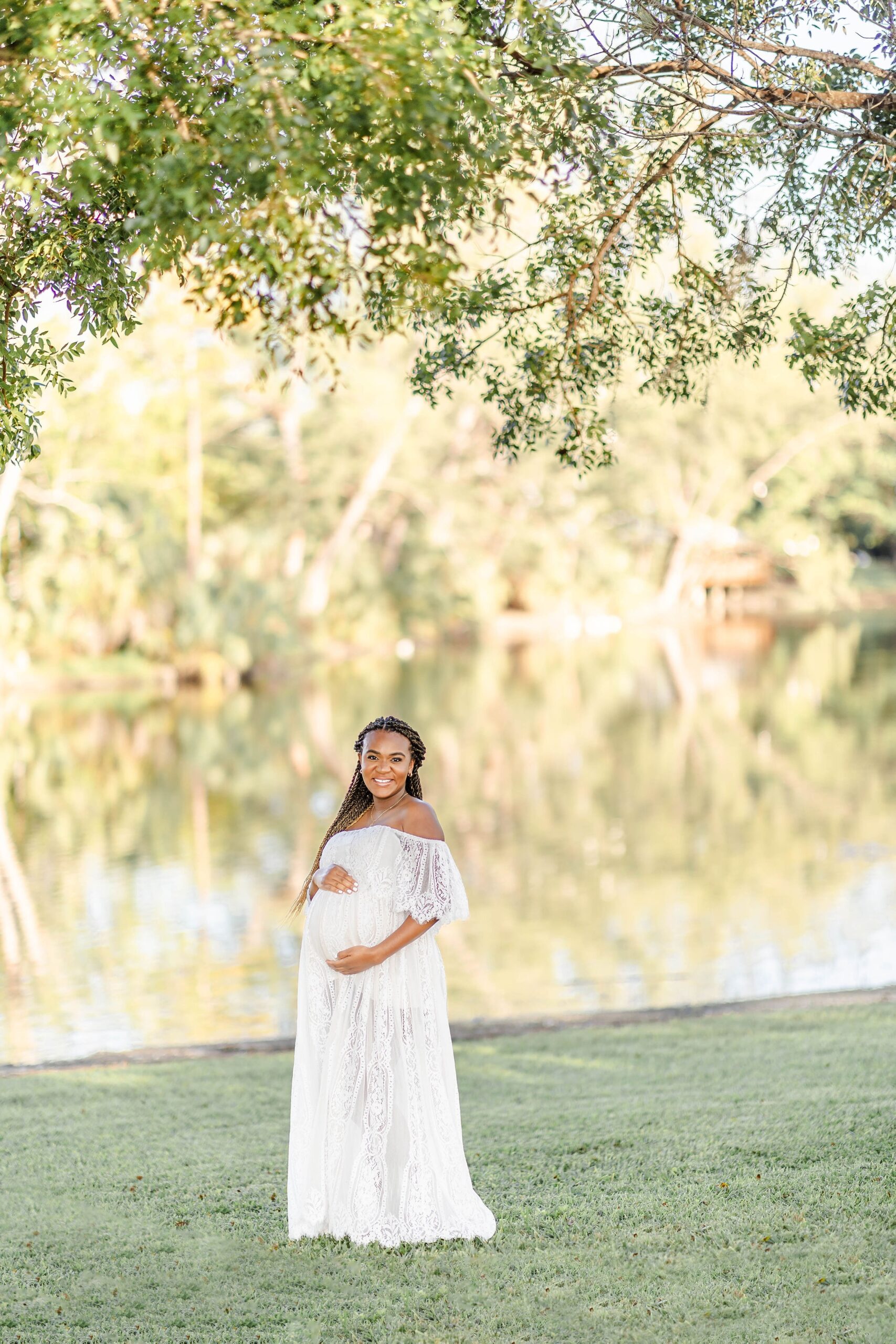 A happy pregnant woman stands by a lake holding her bump in a white lace maternity gown after visiting tlc women's health