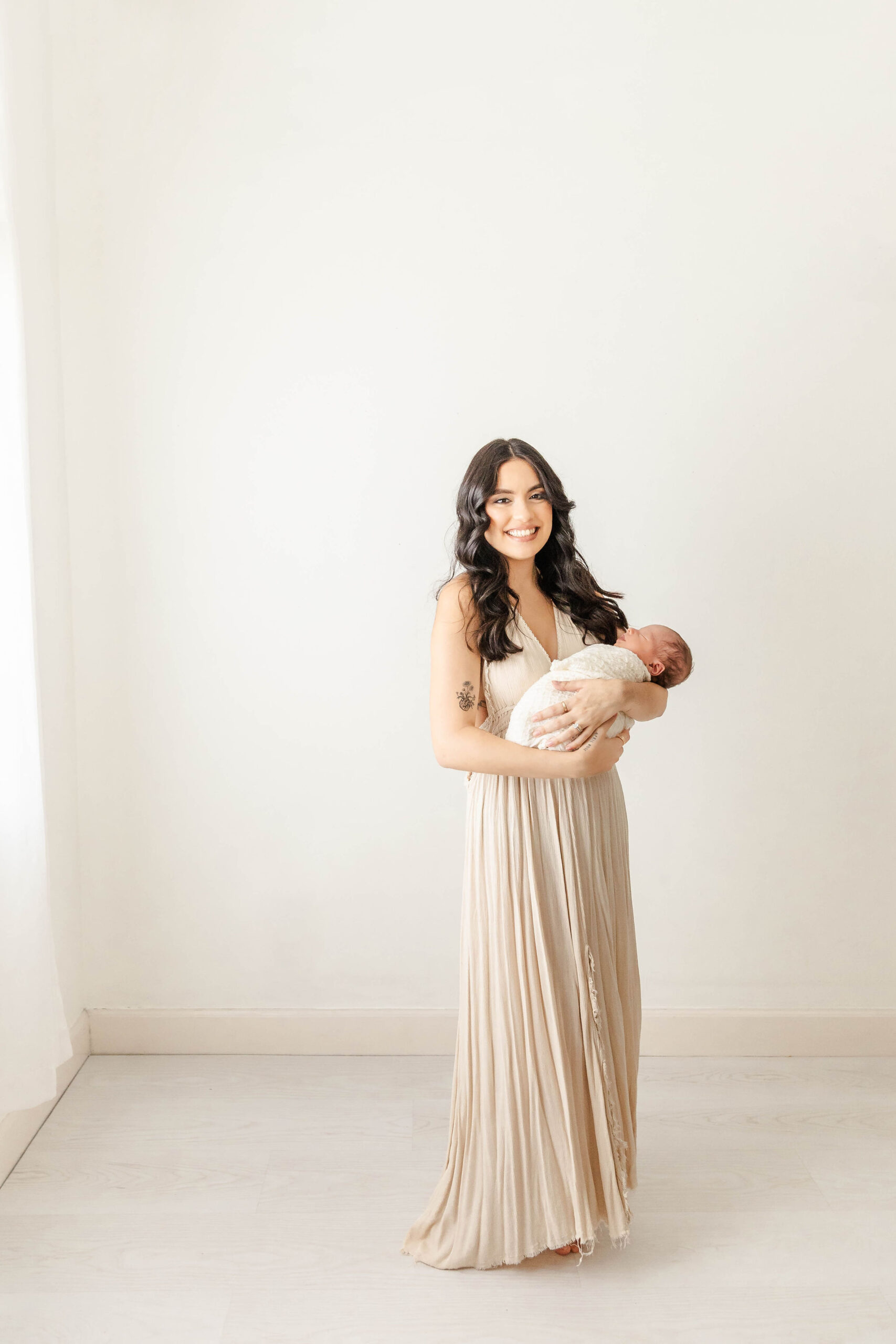 A smiling new mom in a tan gown stands in a studio holding her sleeping newborn after finding infant daycare in fort lauderdale