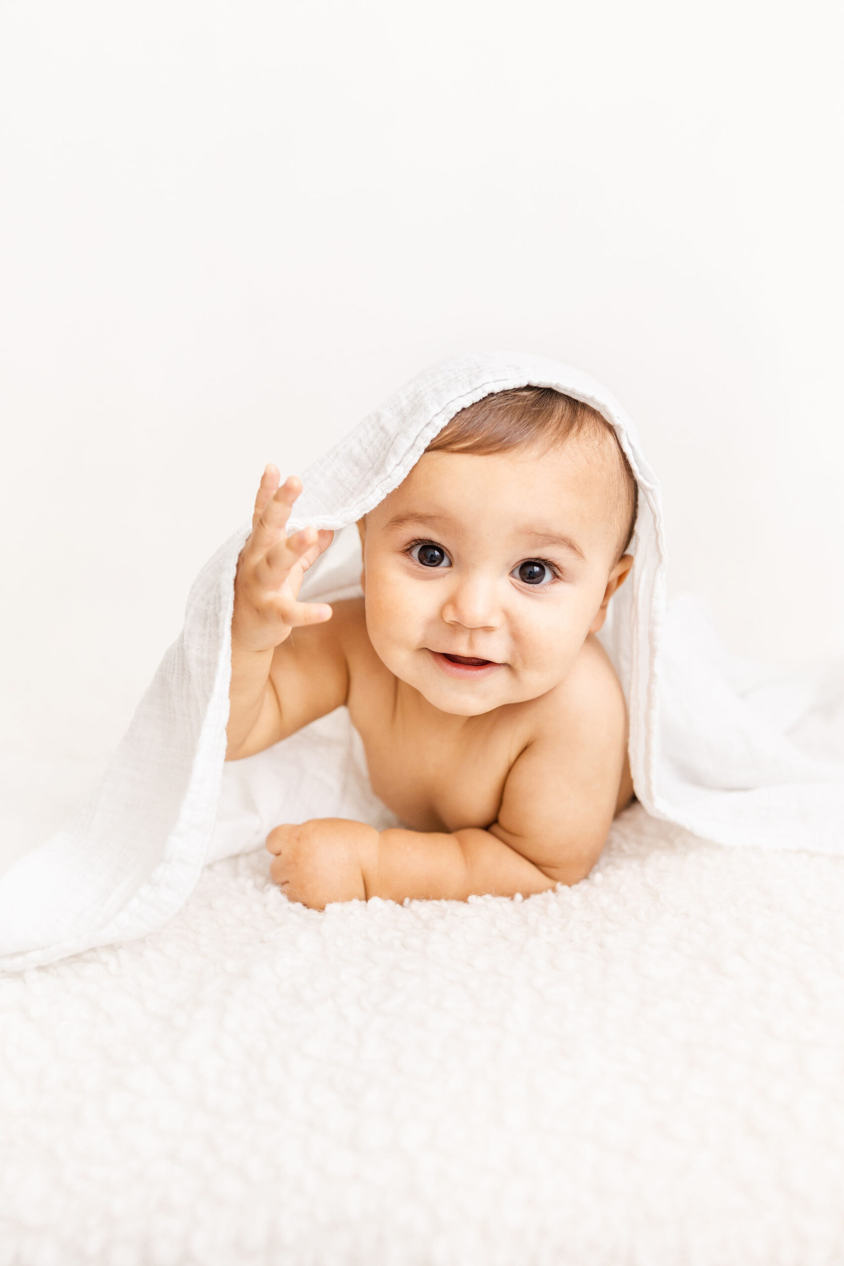 A baby crawls under a blanket on a bed while smiling after using a nanny agency in fort lauderdale