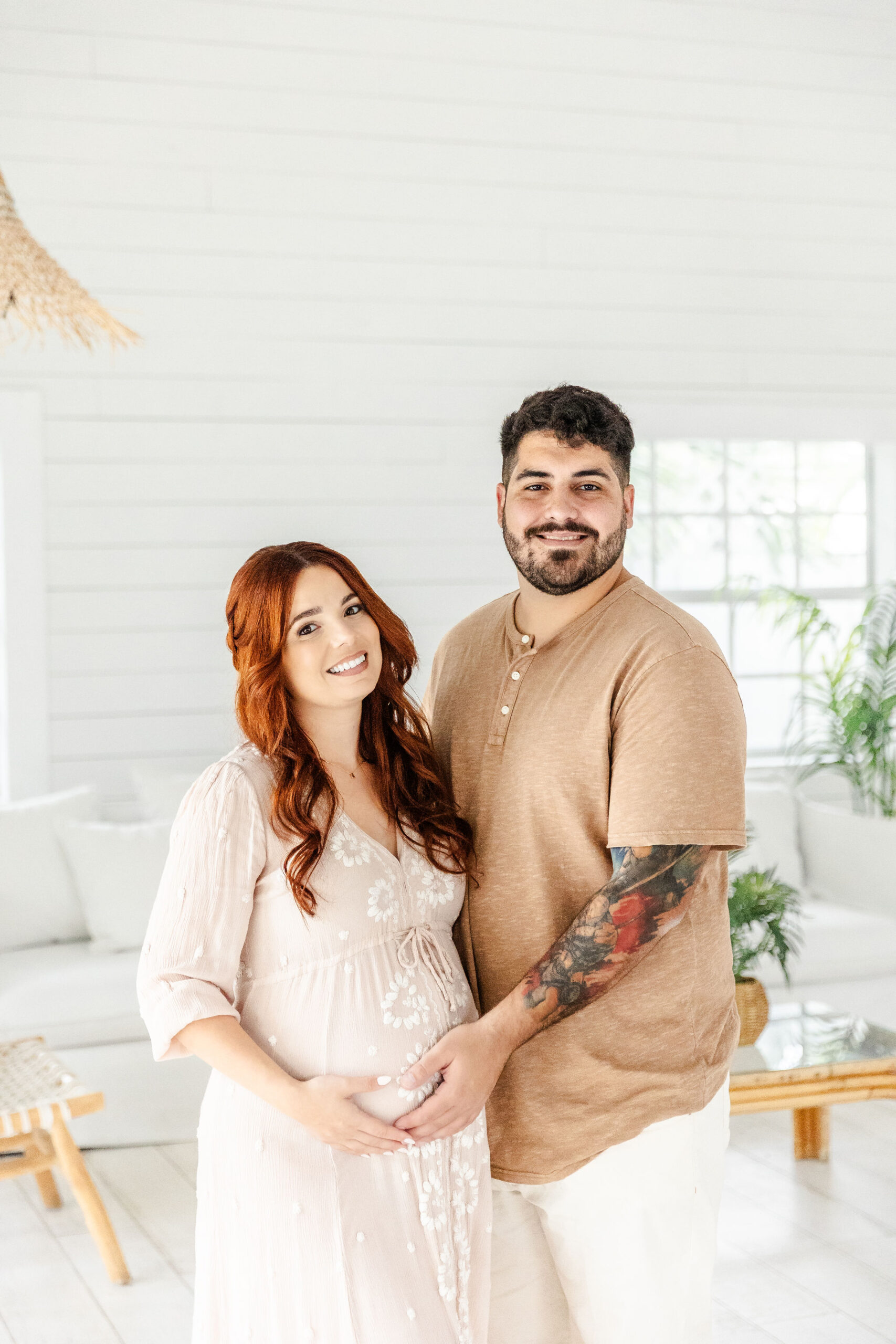 A happy pregnant couple in neutral outfits stand in a white room smiling with hands on bump after a 3d ultrasound in fort lauderdale