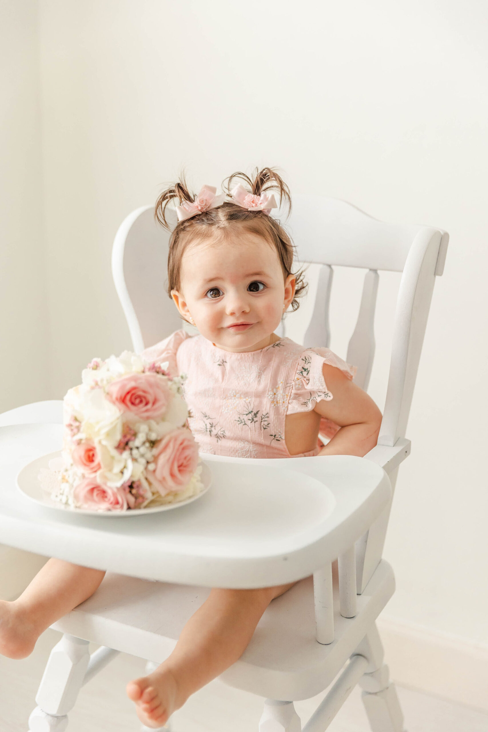 A smiling baby girl in a pink dress sits in a high chair with a flower covered cake