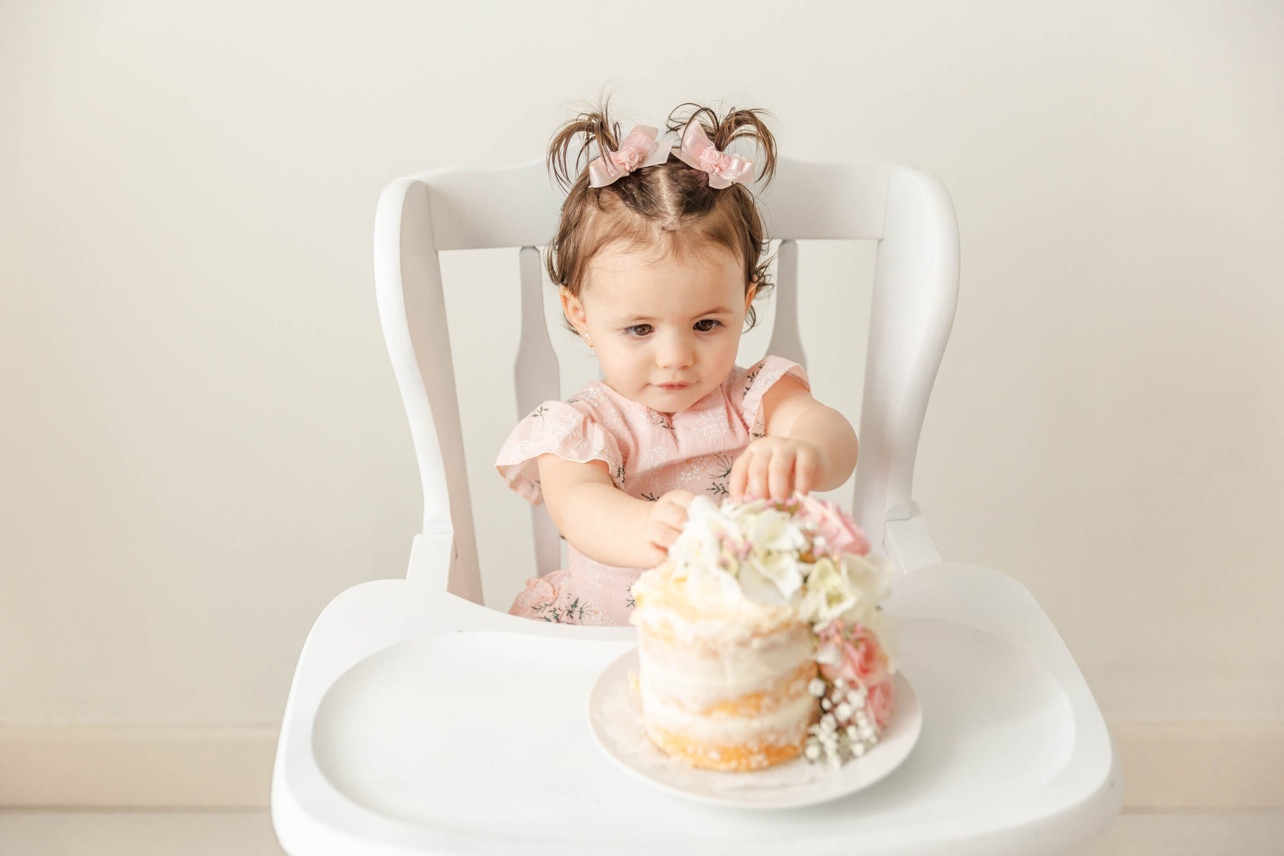 A baby girl in pigtails and a pink dress reaches for a cake in her high chair in a studio after visiting pediatric dentists in fort lauderdale