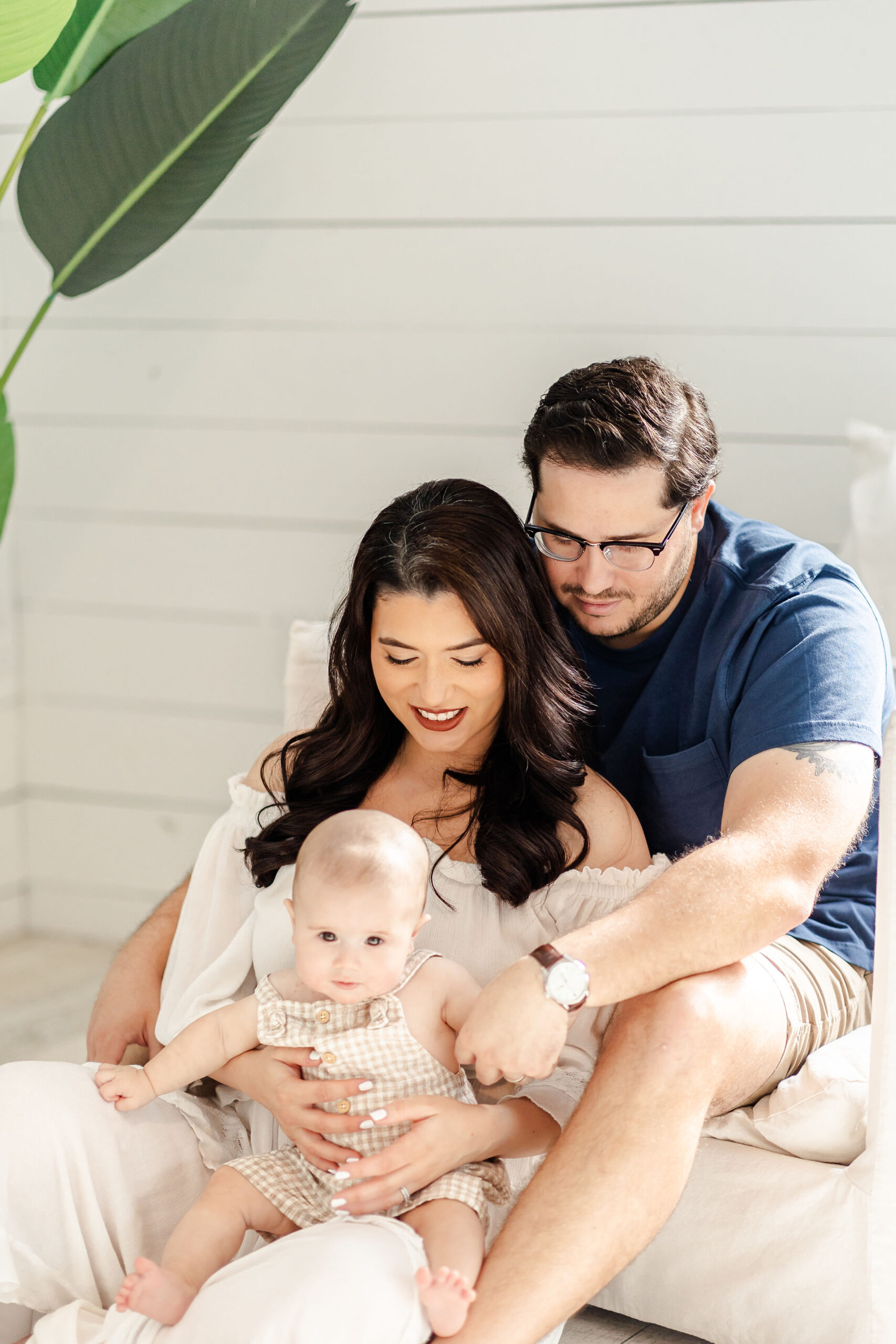 A baby sits in mom's lap as she snuggles into dad's on a chair in a studio after visiting pediatricians in fort lauderdale