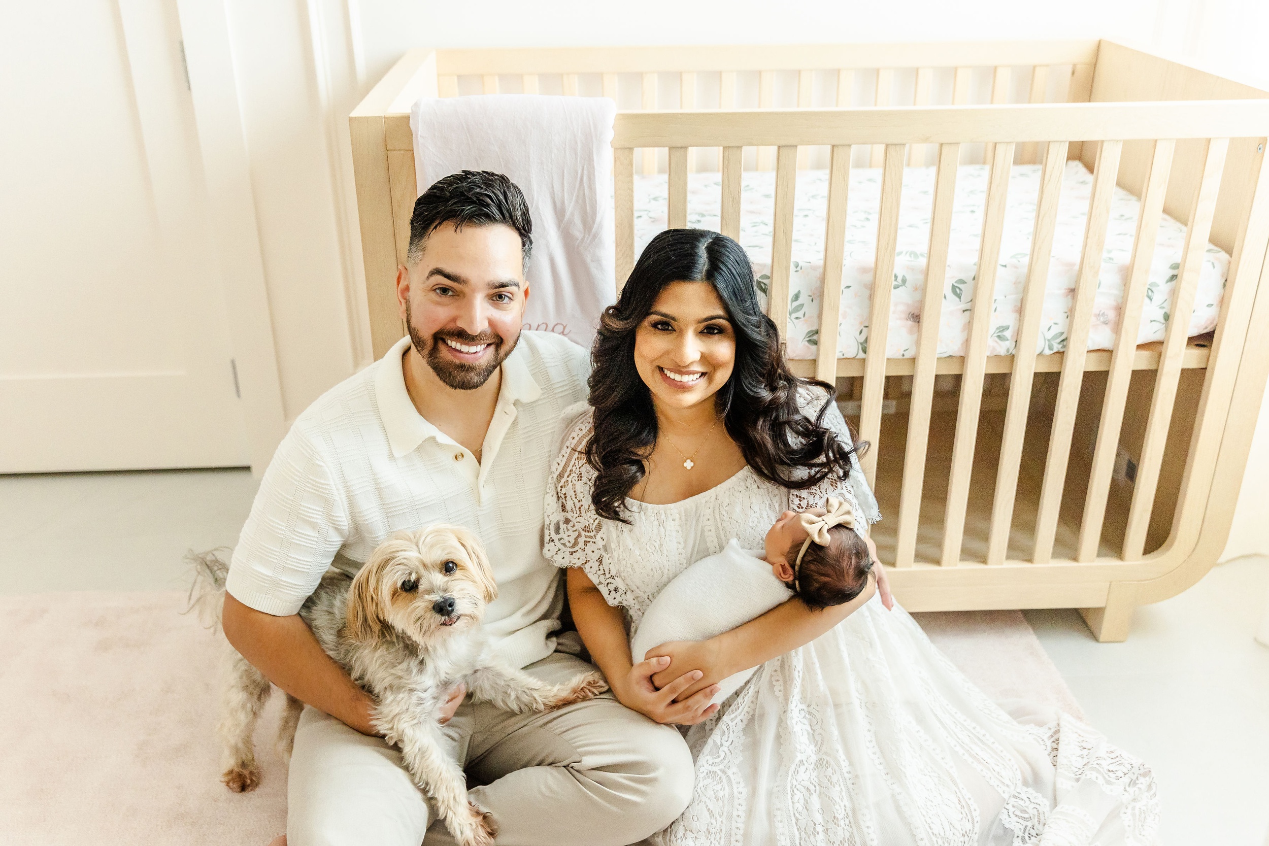 Happy new parents sit leaning on a wooden crib holding their newborn girl and tiny dog in white after meeting a lactation consultant in fort lauderdale