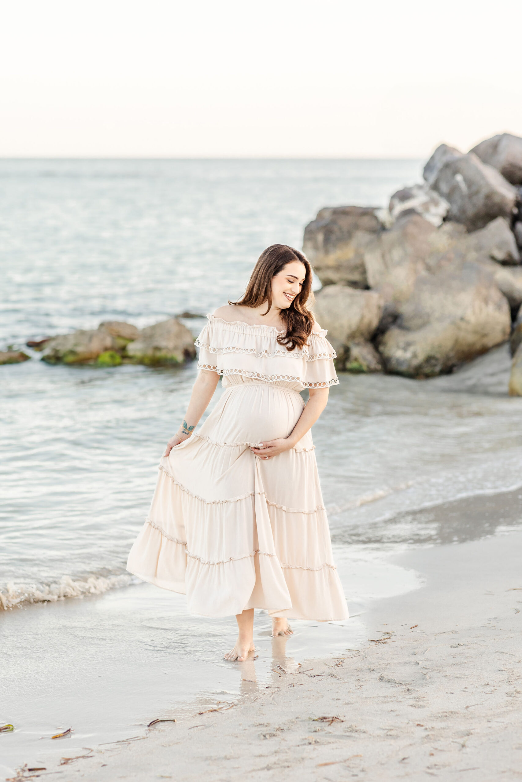 A laughing mother to be in a cream dress dances in the water on the beach