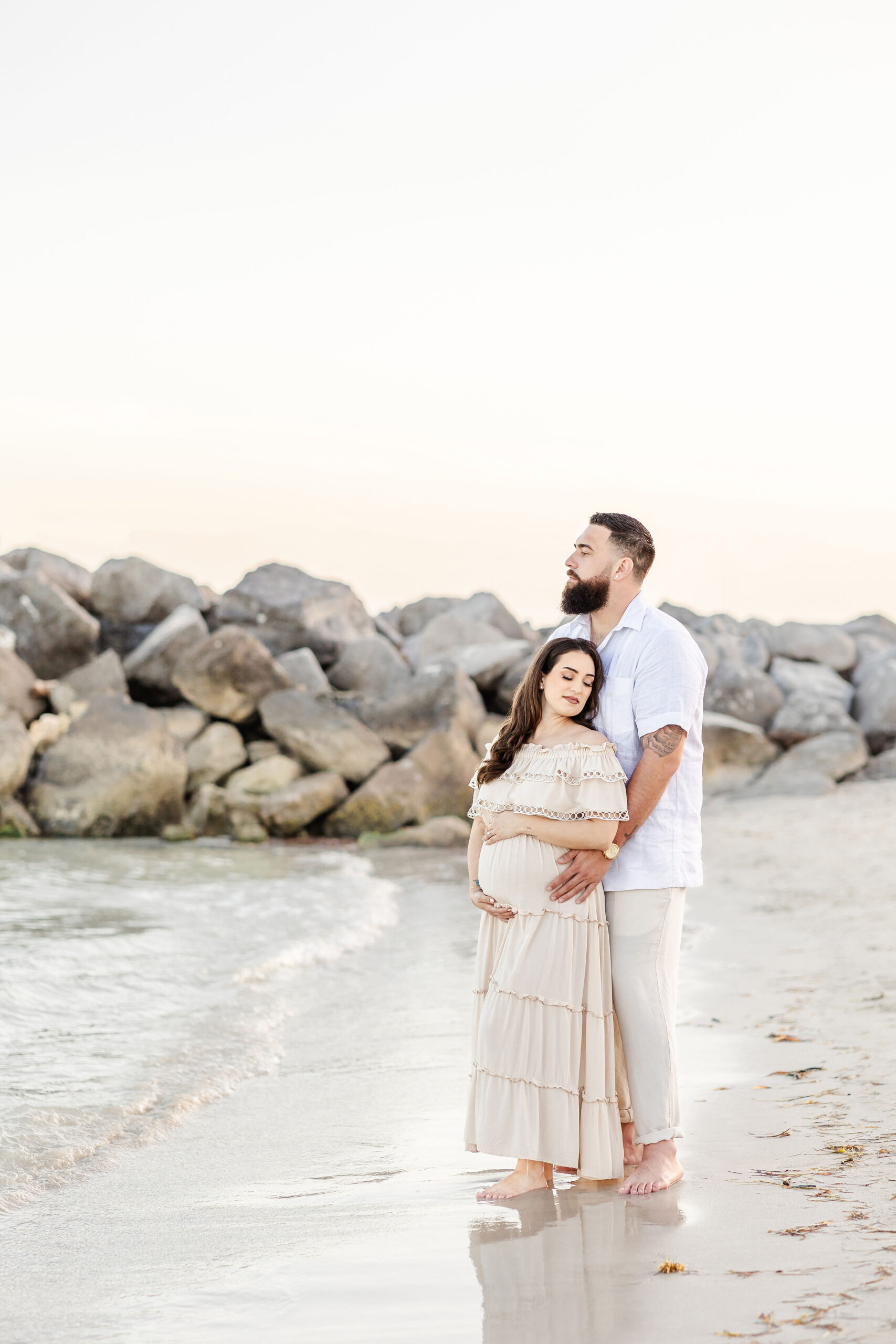 A happy pregnant couple snuggles on a beach at sunset while standing in the water's edge after visiting an obgyn in fort lauderdale