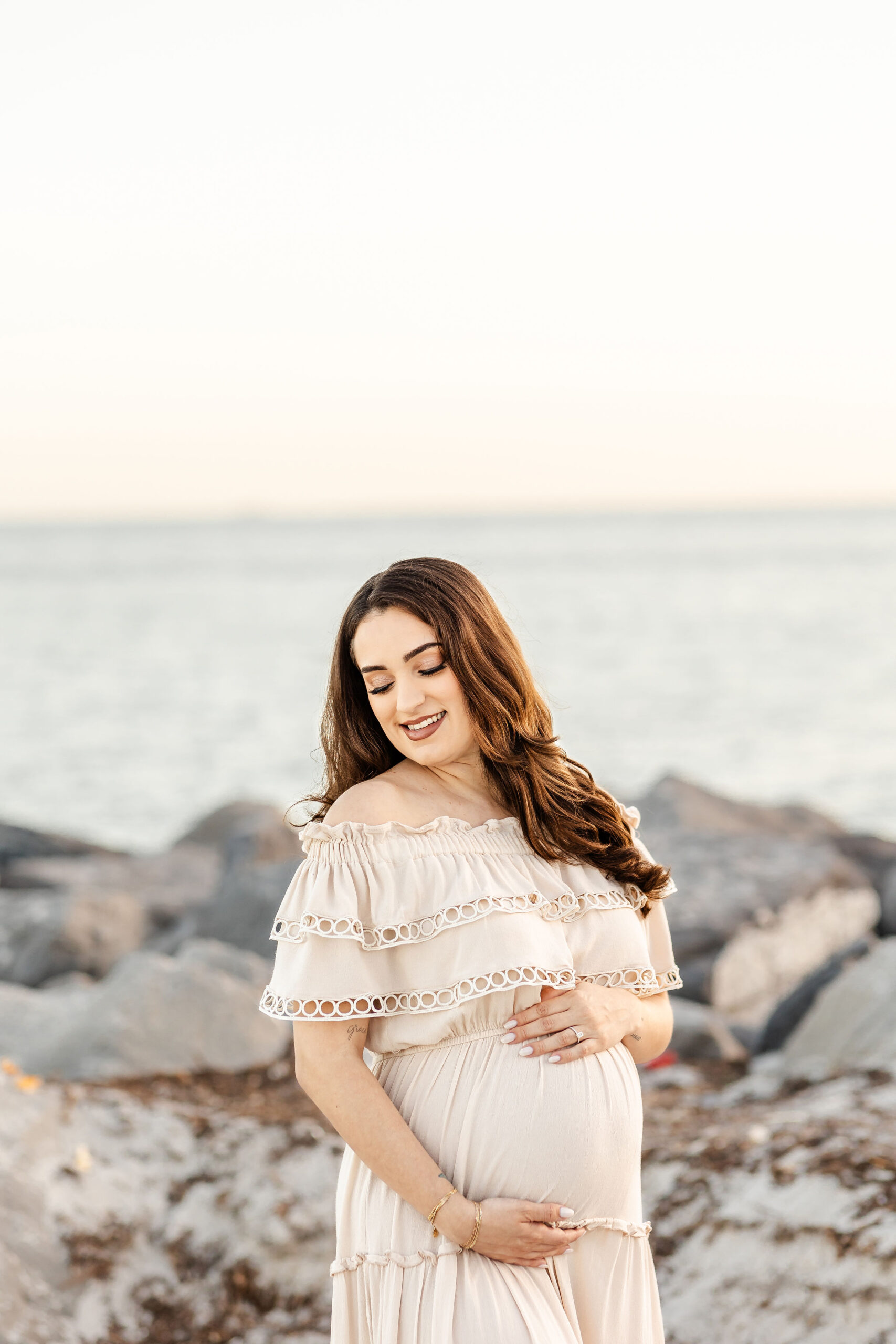 A mother to be in a cream maternity dress smiles down her shoulder on a rocky beach at sunset after finding an obgyn in fort lauderdale
