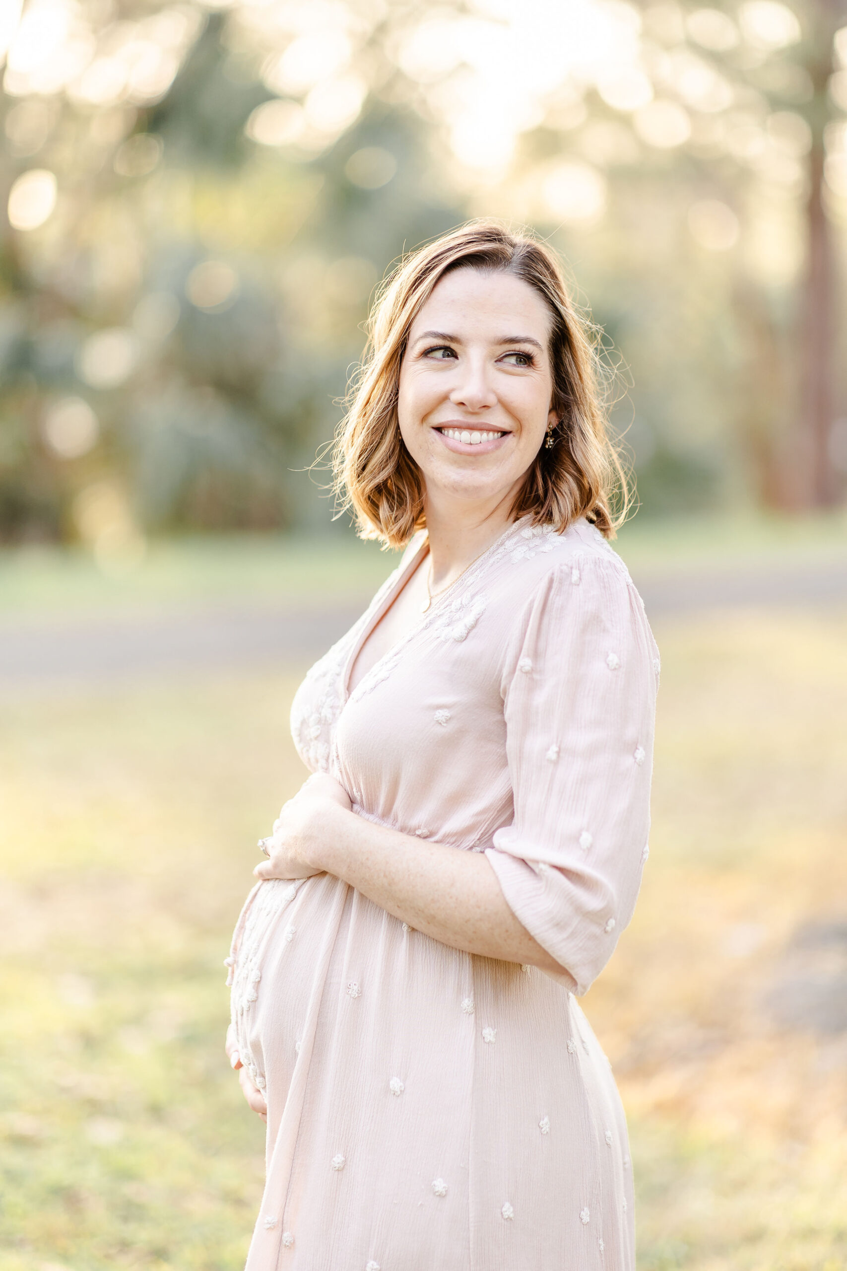 A happy mom to be in a pink maternity gown smiles over her shoulder with hands on her bump at sunset