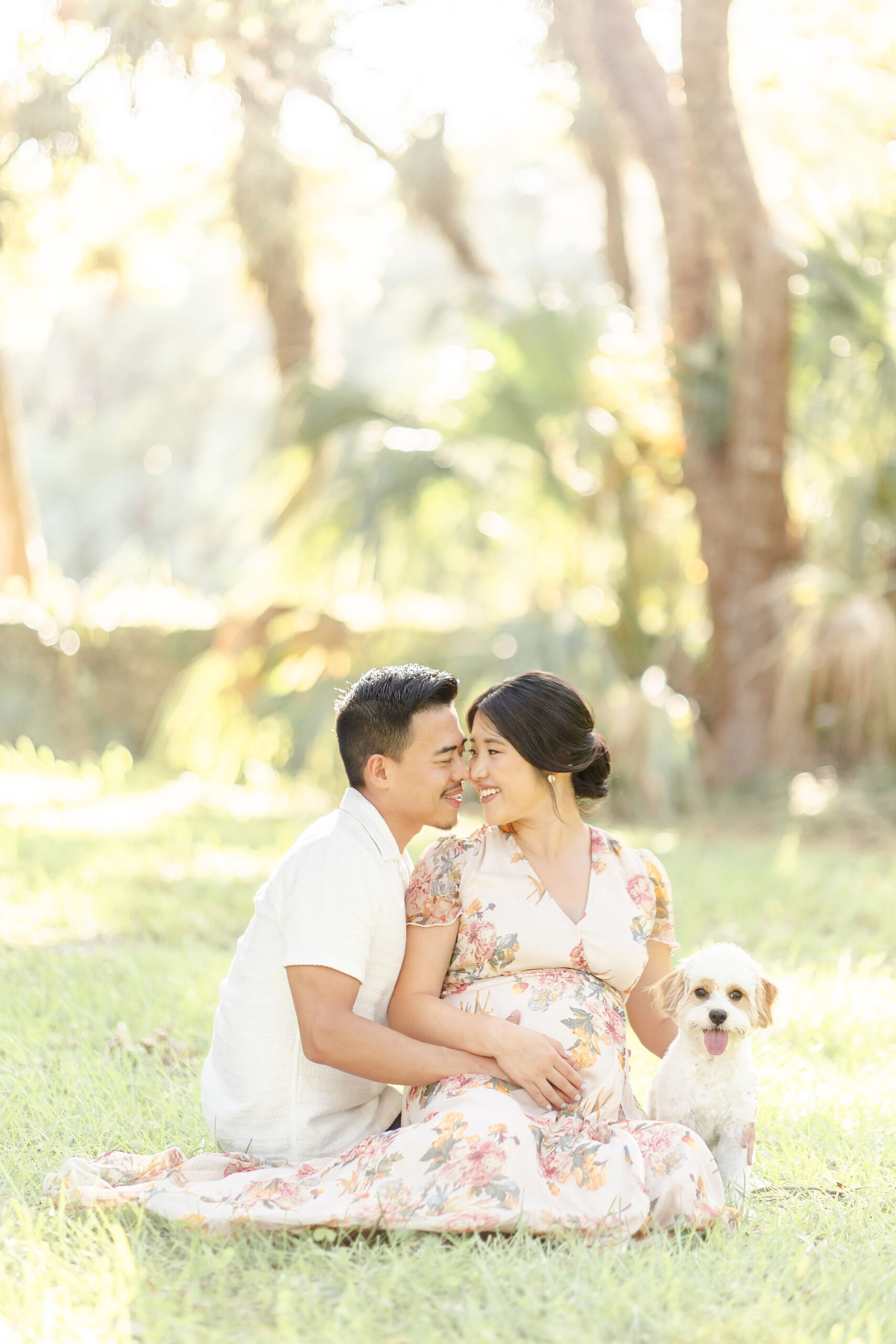 A pregnant woman sits with her small dog in a field while her husband leans in to kiss her