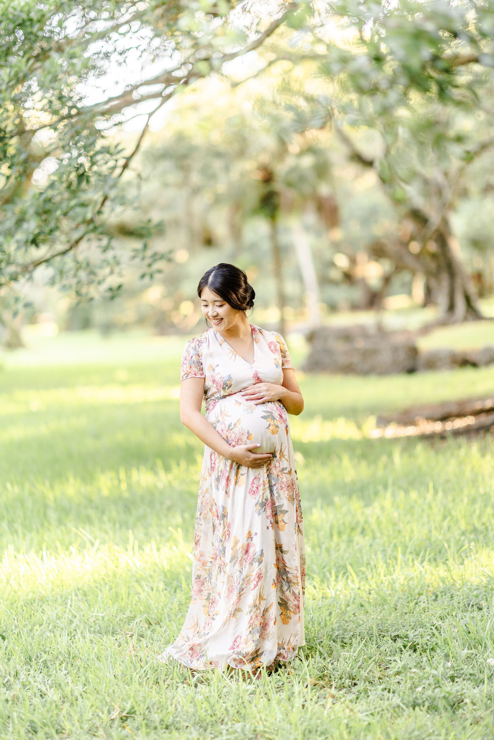 A mom to be in a floral print dress smiles while standing under a tree after some prenatal yoga in fort lauderdale