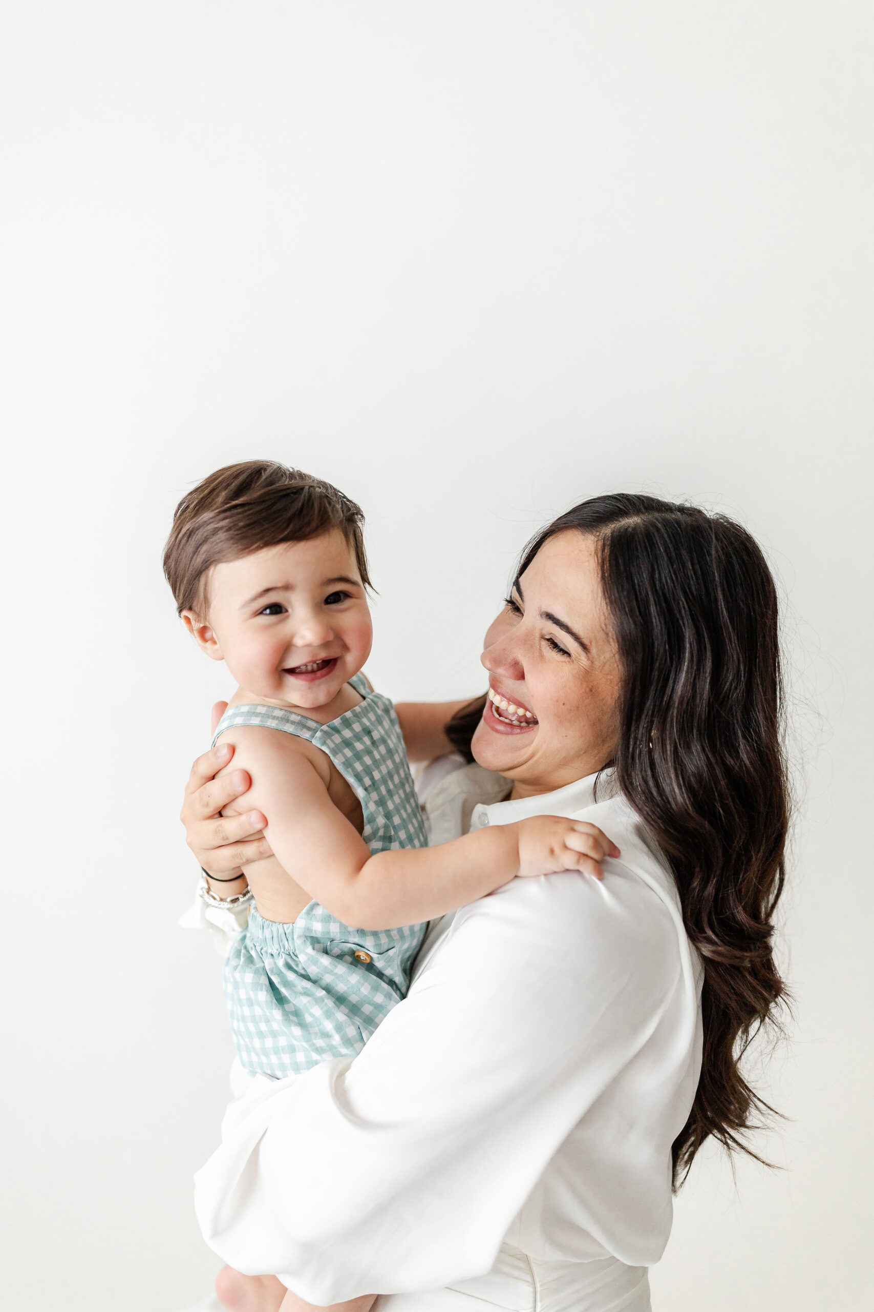 A mother in white laughs with her giggling baby in her arms in a studio