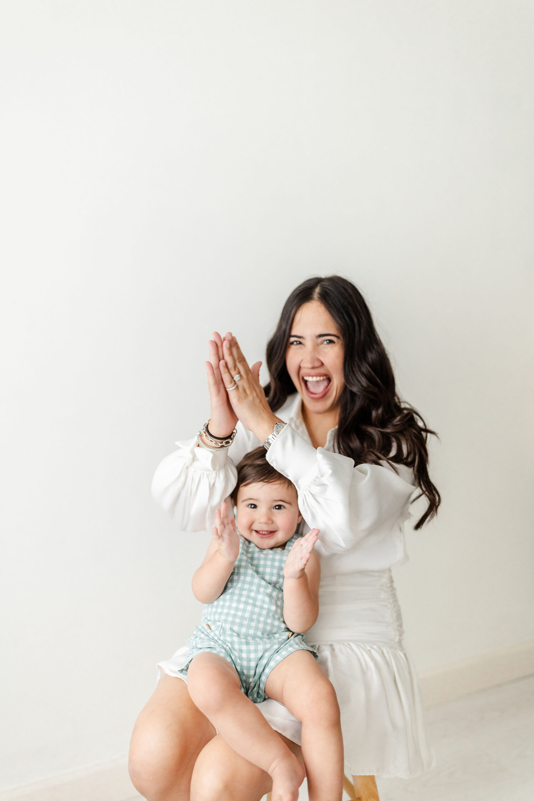 A mom in a white dress claps with her toddler sitting in her lap in a green onesie after some mommy and me classes in fort lauderdale