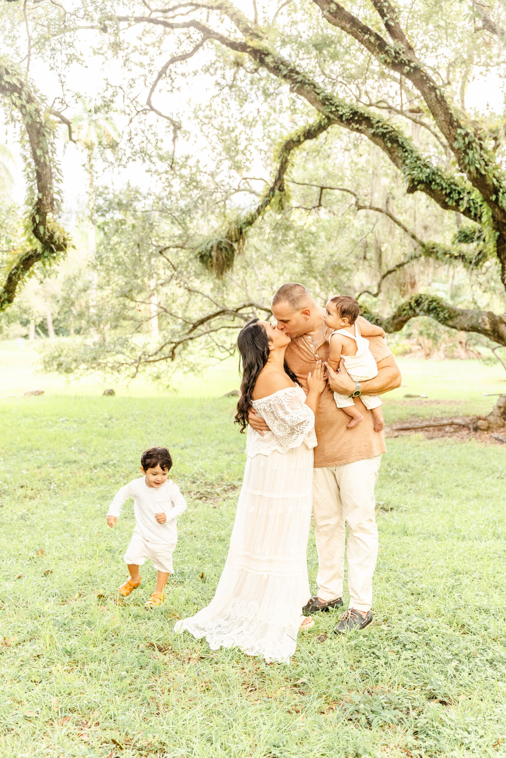 Happy mom and dad kiss while their toddler runs around them under old live oaks in a park and holding infant after parenting classes in Fort Lauderdale