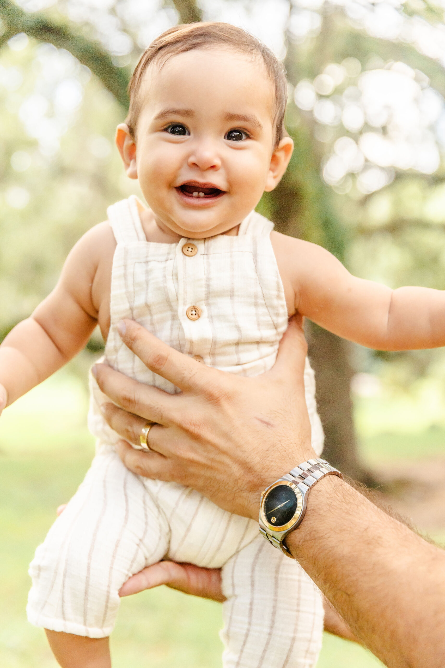 A happy baby is lifted by dad in a white onesie in a park with a big smile after some parenting classes in Fort Lauderdale