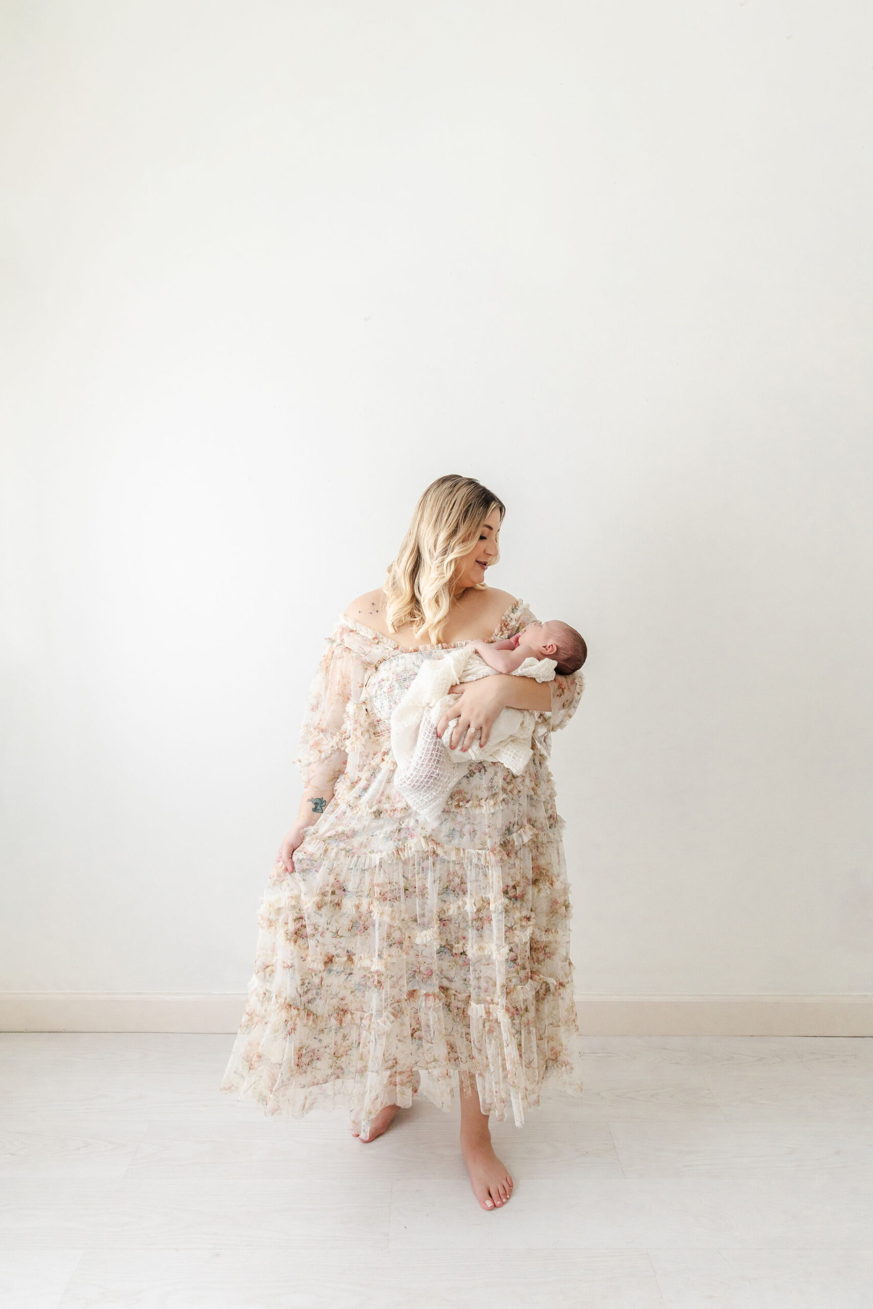 A happy new mom in a floral dress stands in a studio holding her sleeping newborn in one arm before some pelvic floor therapy in fort lauderdale