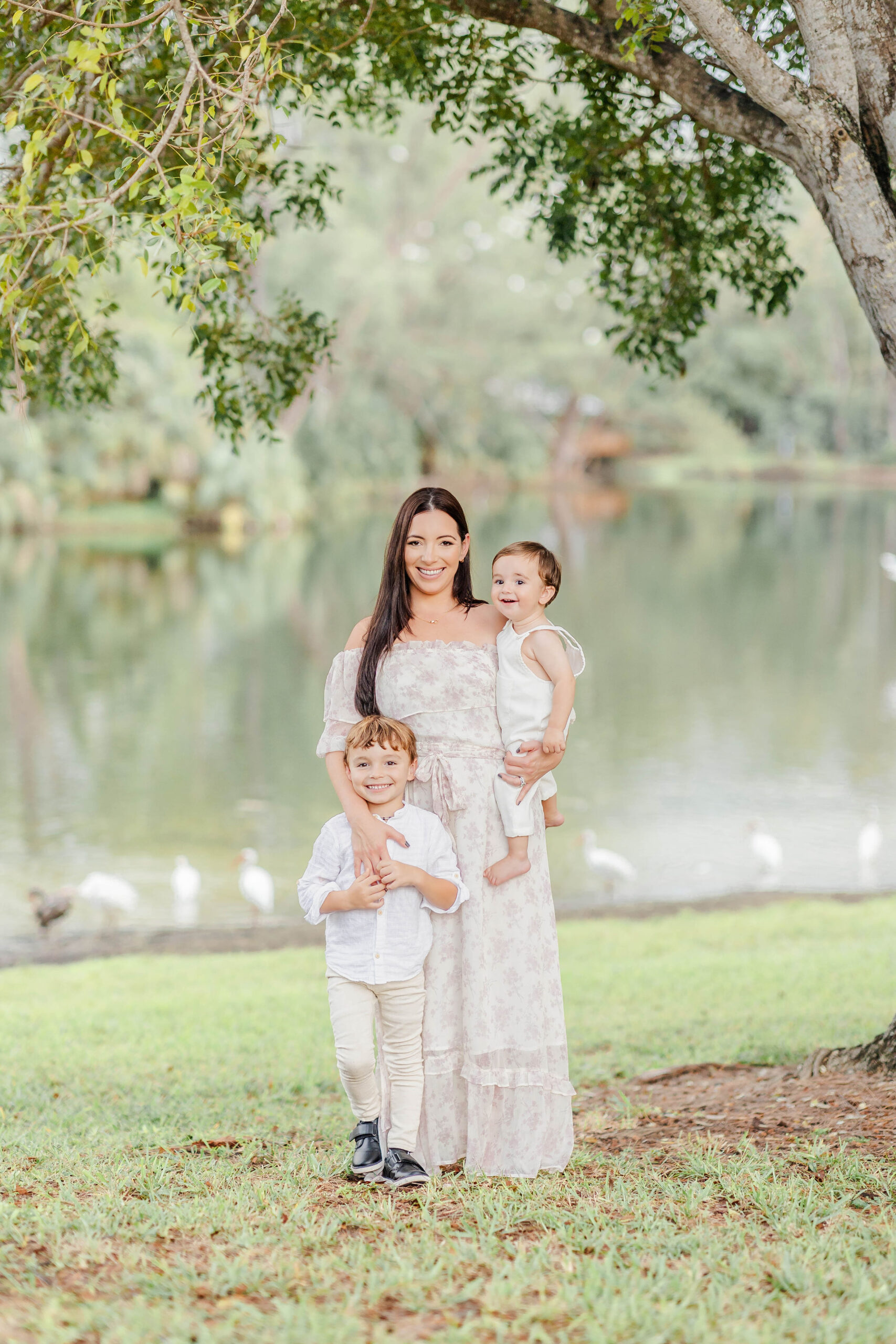 A happy mom in a white dress smiles with her toddler and infant sons by a lakeshore under a tree