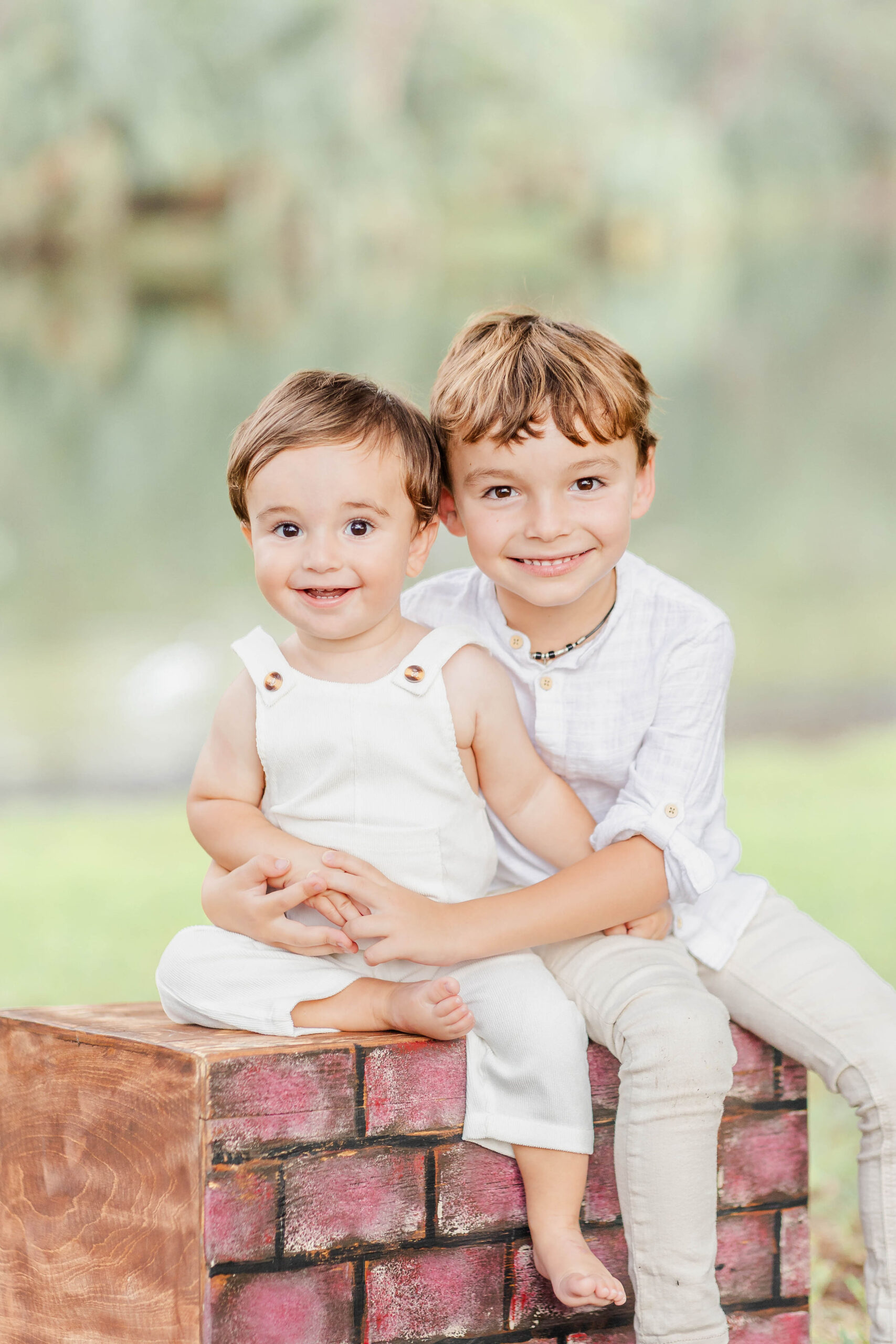 A toddler boy hugs his baby sibling on a box in a park as they both smile in white after visiting preschools in Fort Lauderdale