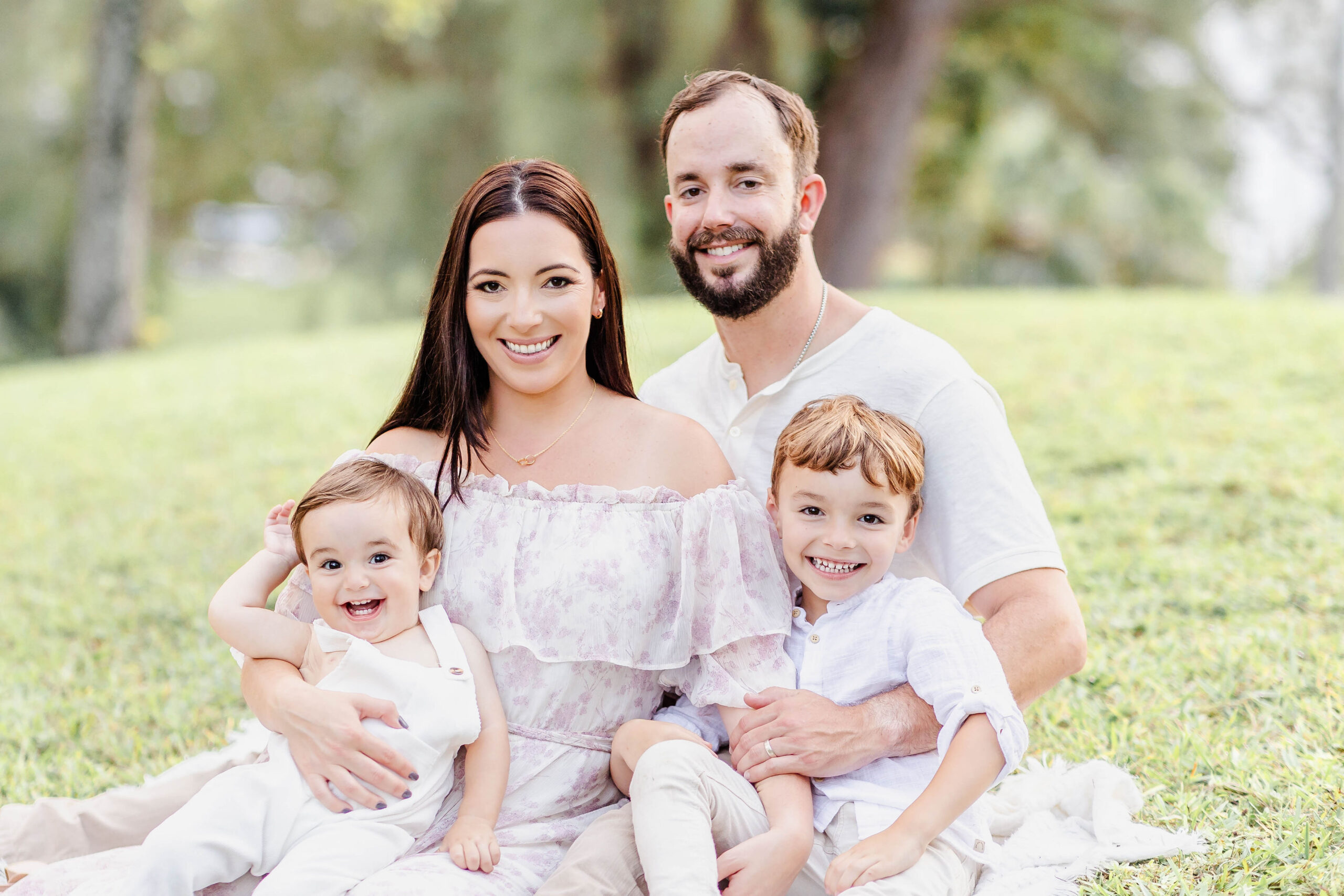 Happy mom and dad smile with their giggling toddlers in their laps all in white in a park lawn after finding preschools in Fort Lauderdale