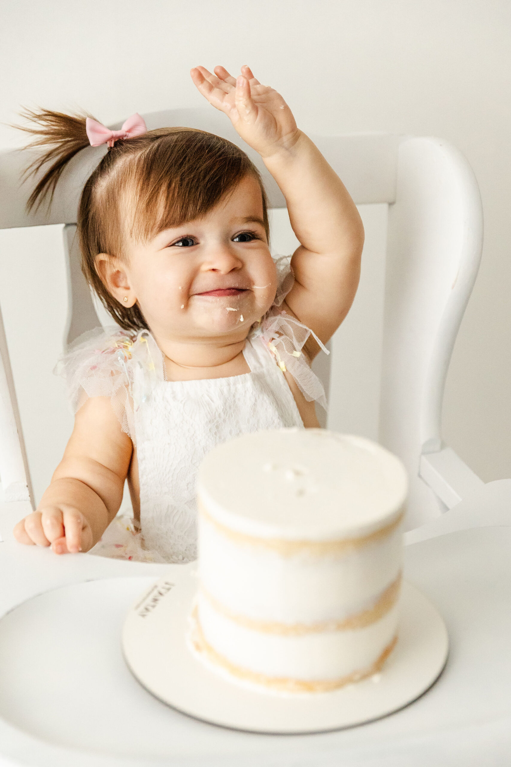 A happy infant girl sits in a high chair raising her hand while eating her birthday cake