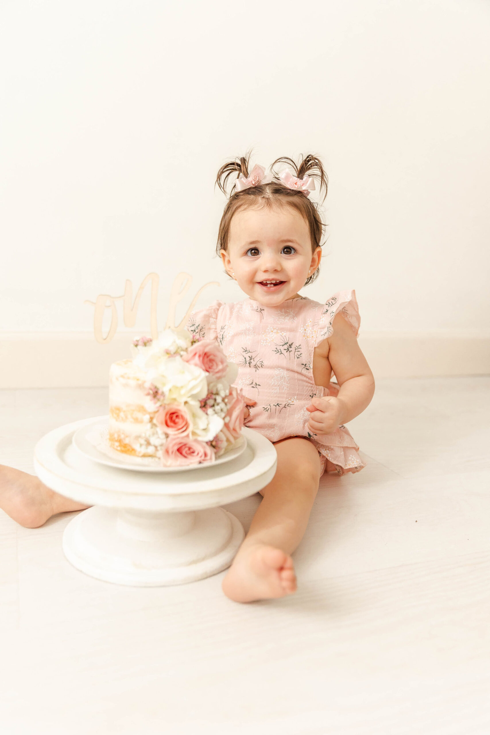 A happy baby girl in a pink dress smiles while sitting behind her first birthday cake on the floor before visiting birthday party places in fort lauderdale