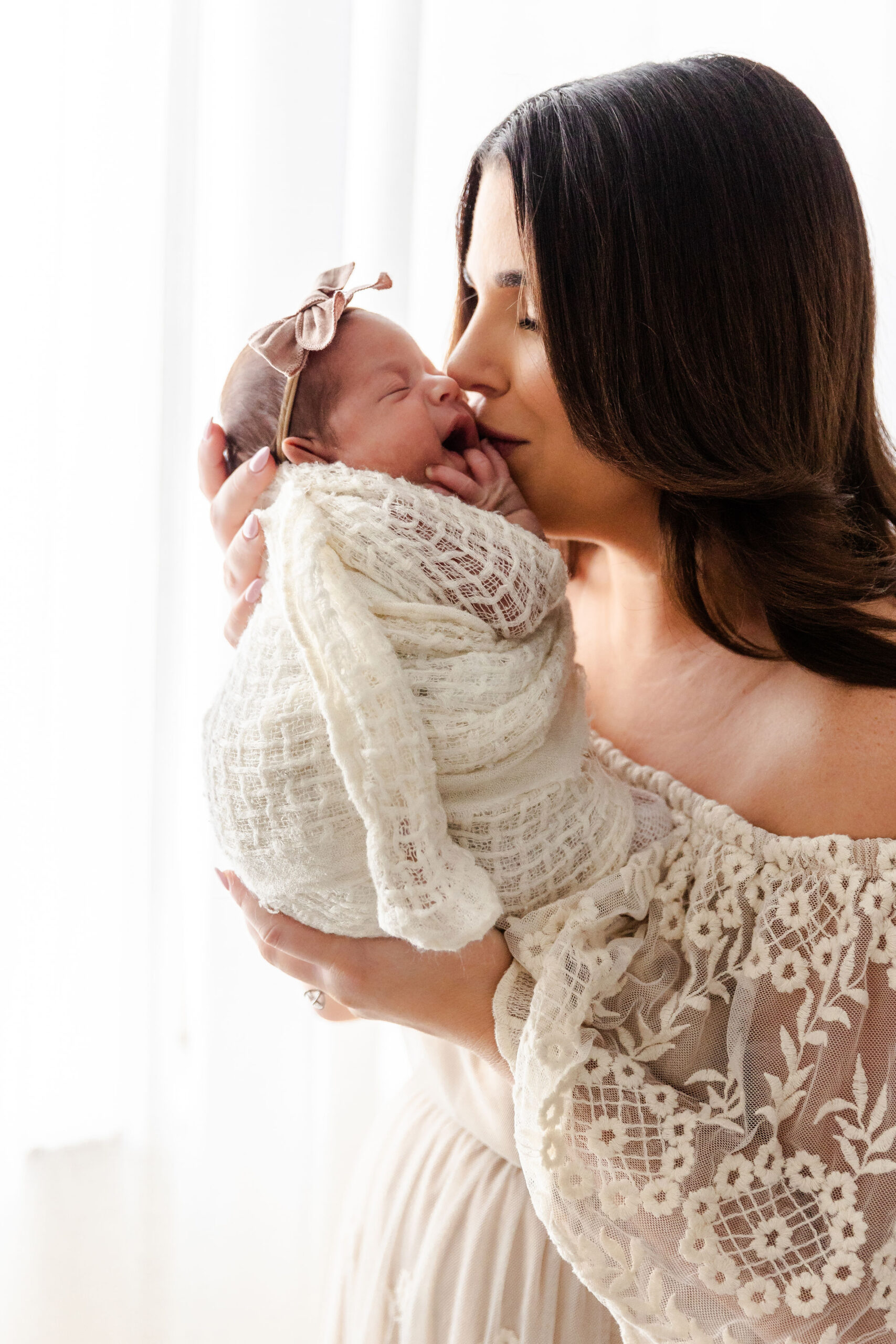 A newborn baby sleeps cradled in mom's hands and a swaddle as mom kisses her on the cheek while standing in a window before some infant swim lessons in fort lauderdale