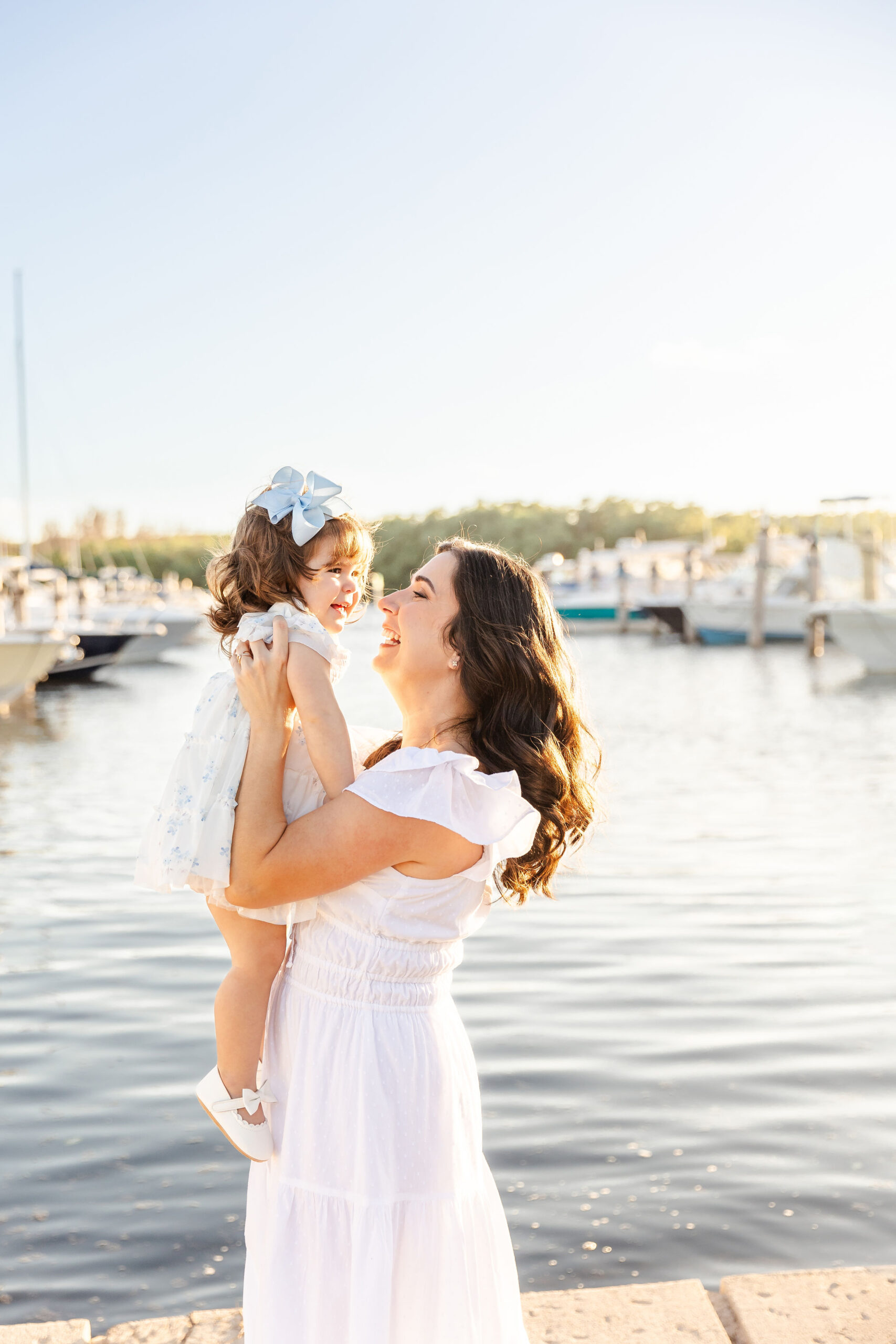 A joyful mother in a white dress lifts her toddler daughter in a white and blue dress while walking in a marina at sunset