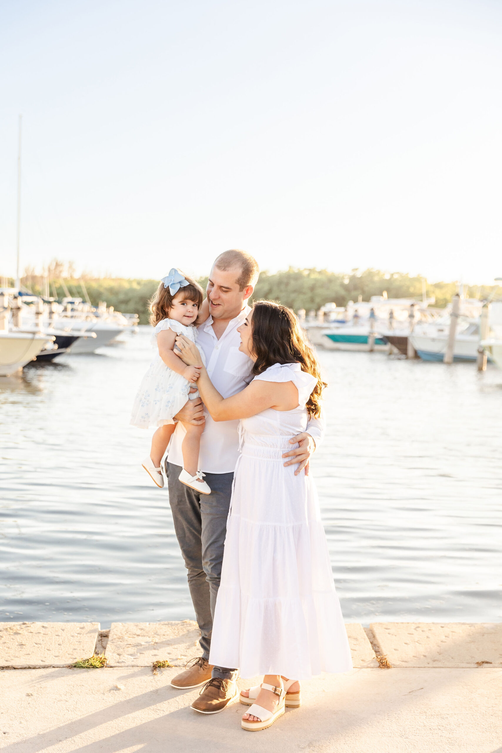A mom and dad walk in the marina while tickling their toddler daughter in a white dress in dad's arms after visiting montessori schools in fort lauderdale