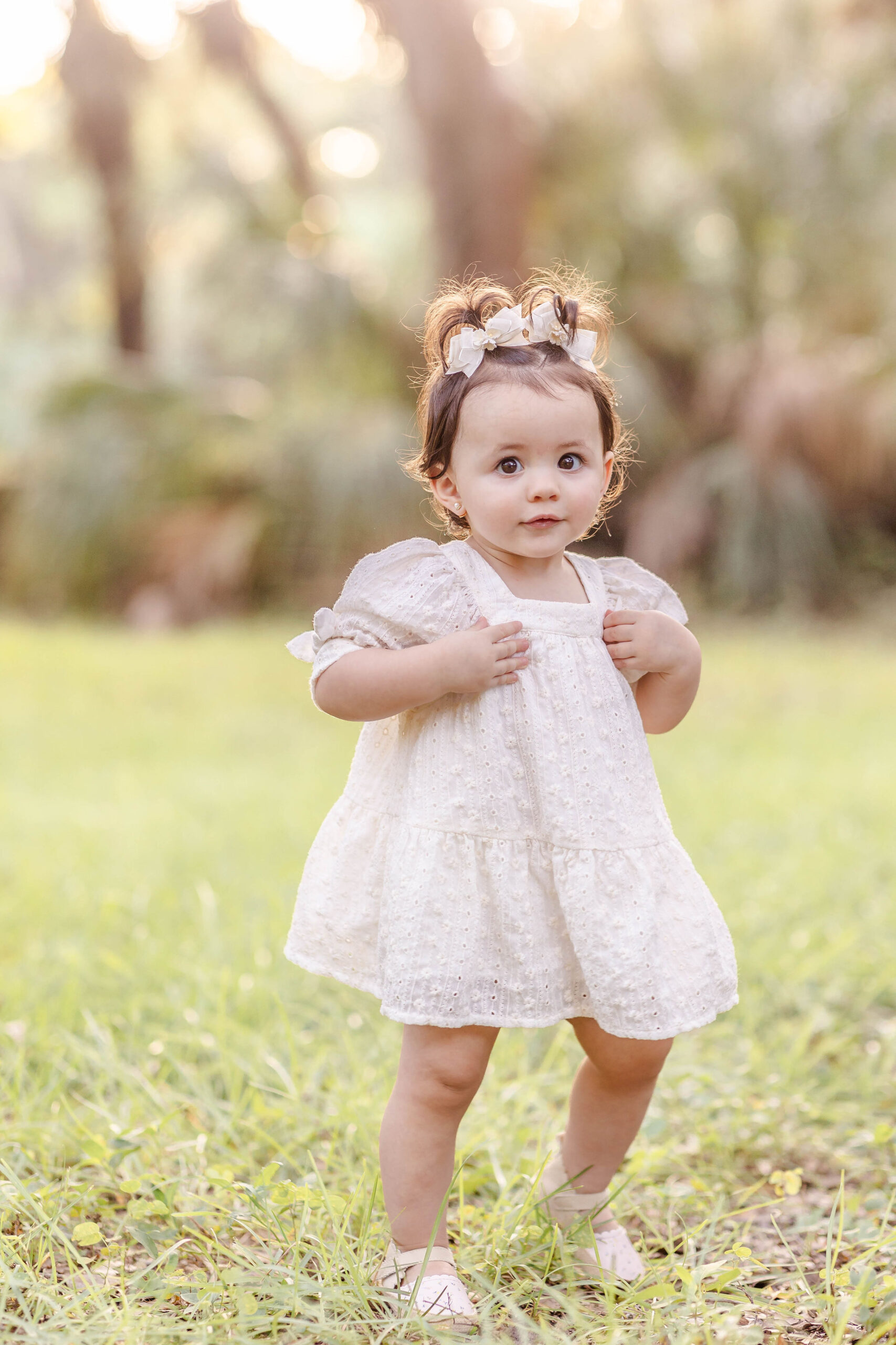 An infant girl in a white dress smiles while exploring a park at sunset