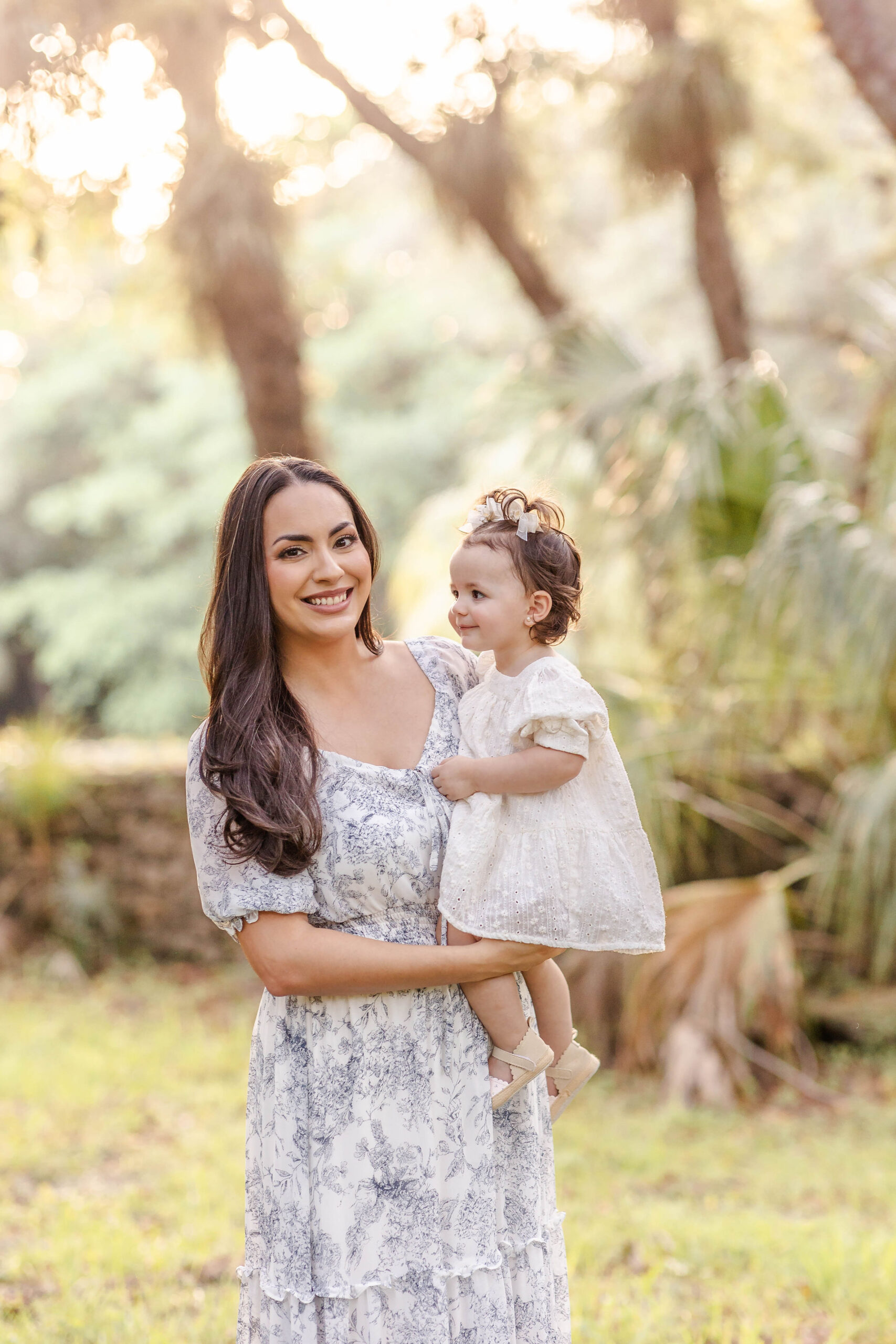 A smiling mom in a floral dress stands in a park at sunset holding her toddler on her hip in a white dress before exploring private preschools in fort lauderdale