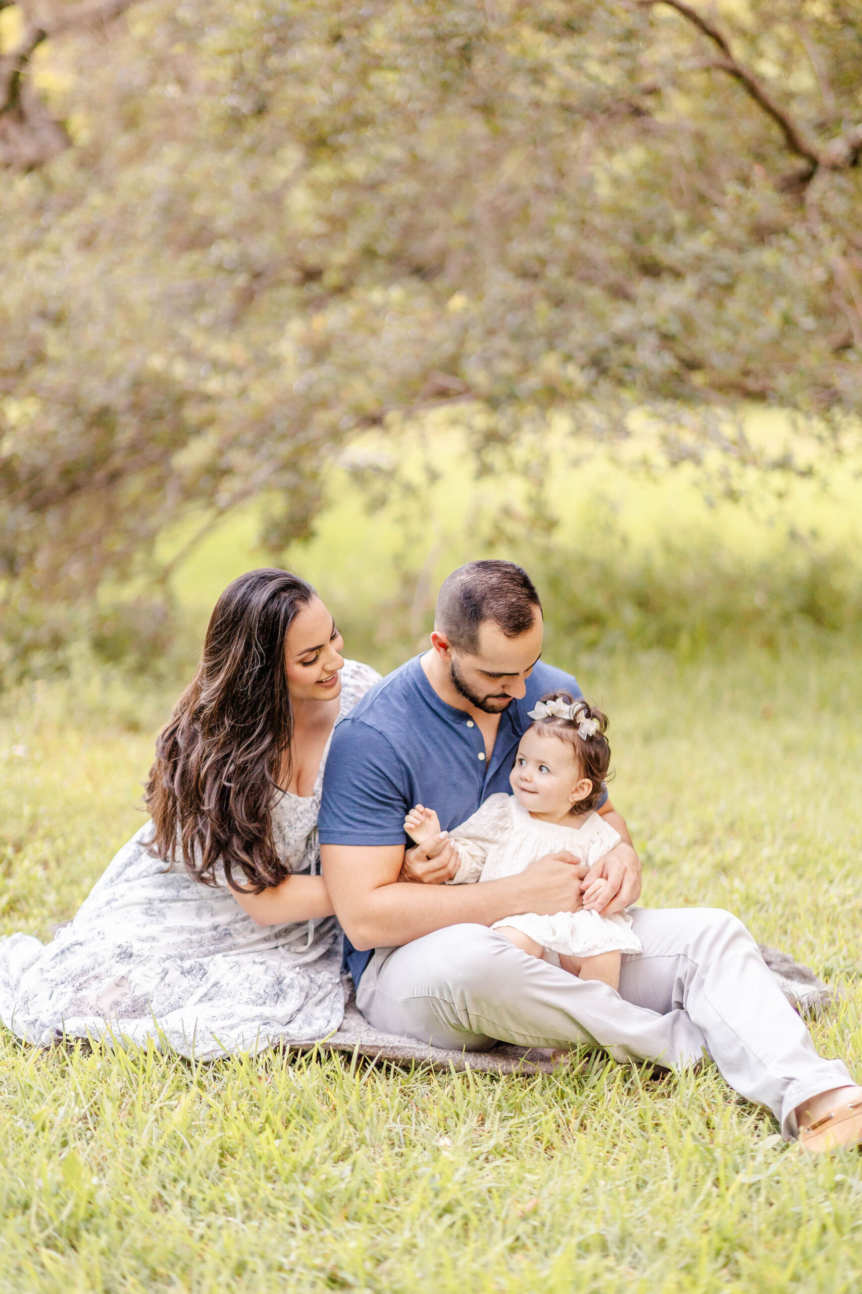 A baby girl sits in dad's lap as mom snuggles on them in a park lawn before visiting private preschools in fort lauderdale