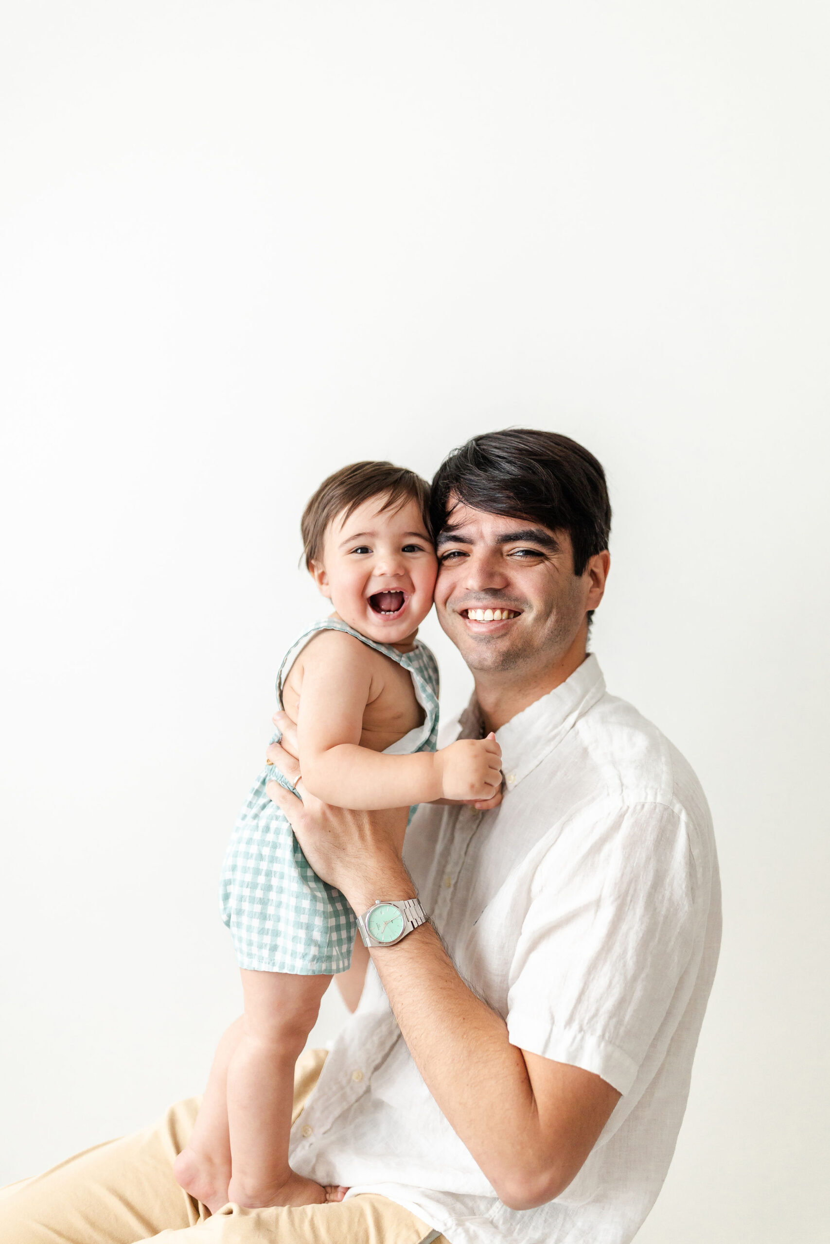 A baby in green plaid overalls laughs while standing in dad's lap and cheek to cheek with him