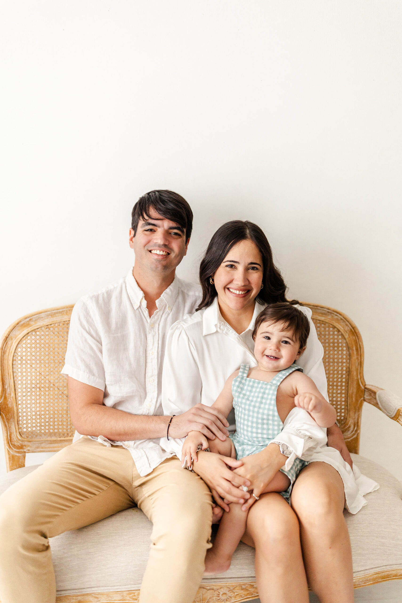 Happy mom and dad sit on a bench in a studio with their smiling infant in mom's lap after visiting toy stores in fort lauderdale