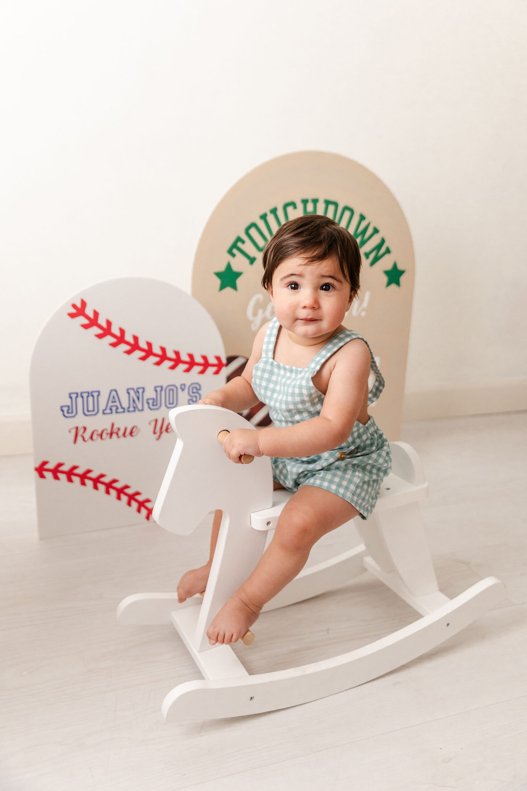 A baby rides a rocking horse in a studio with sports signs behind it in green overalls before visiting indoor playgrounds in fort lauderdale