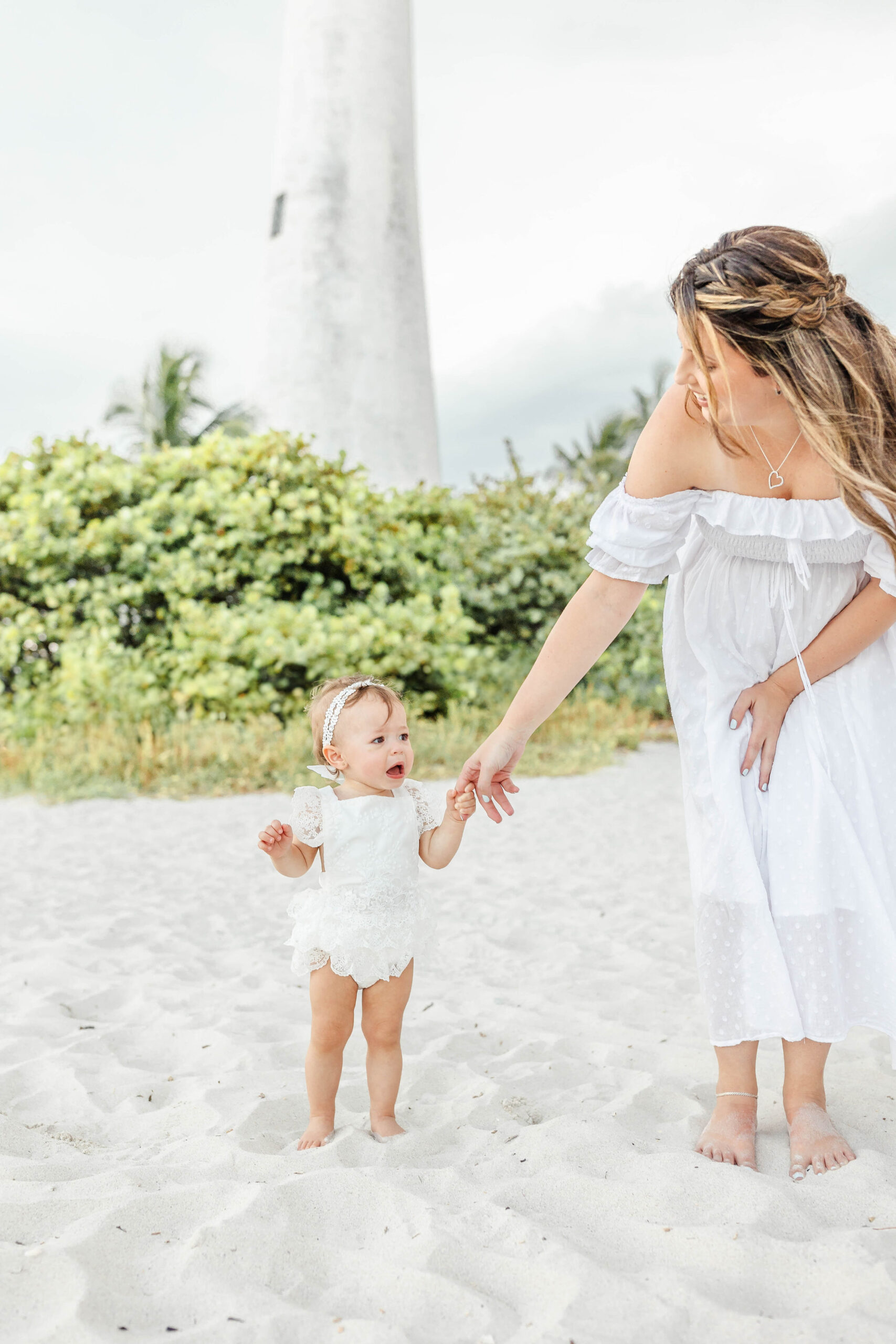 A mom in a white dress helps her toddler walk on the beach with one finger
