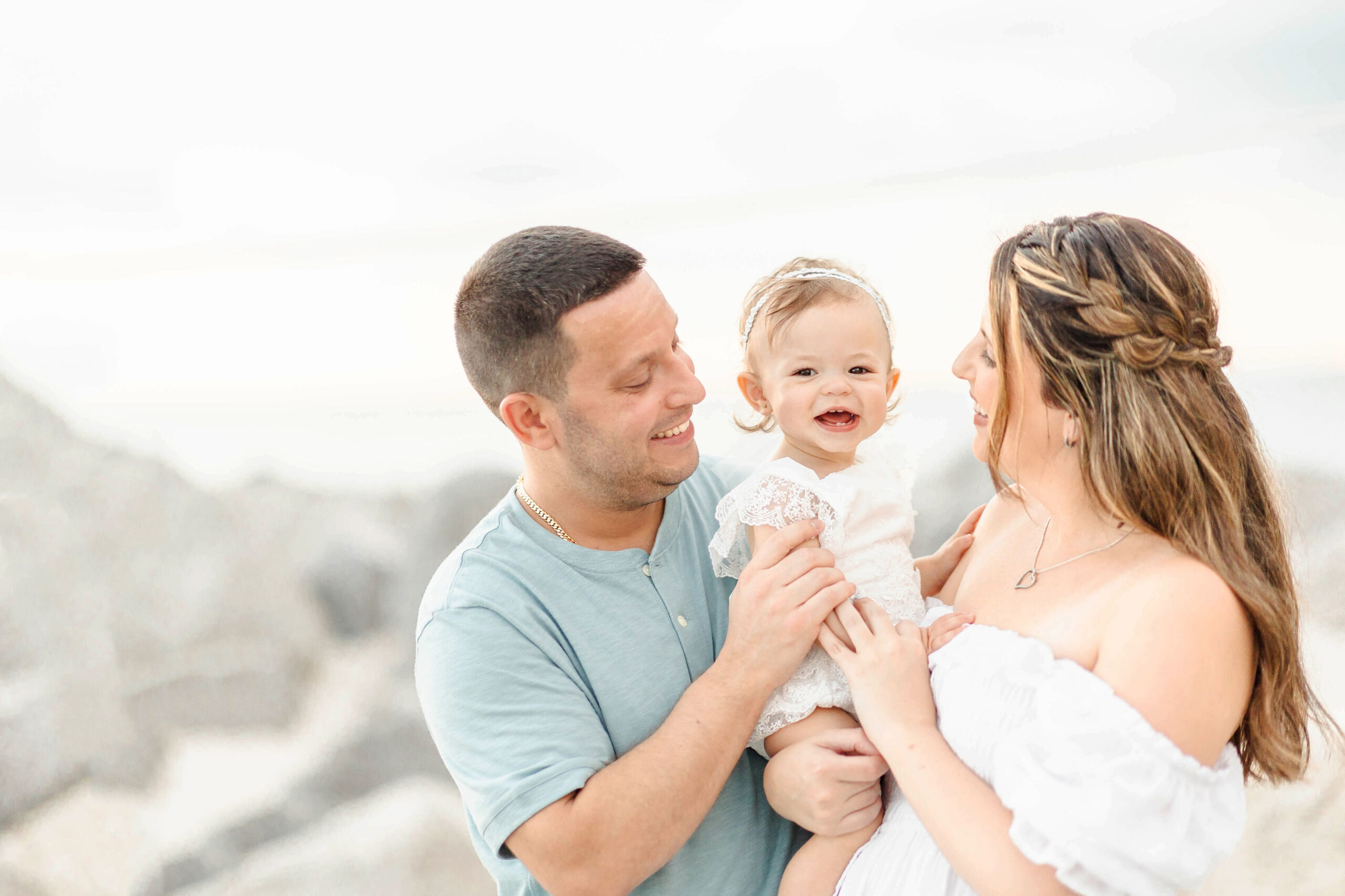 A happy infant in white sits in mom and dad's arms on a beach before visiting splash pads in Fort Lauderdale