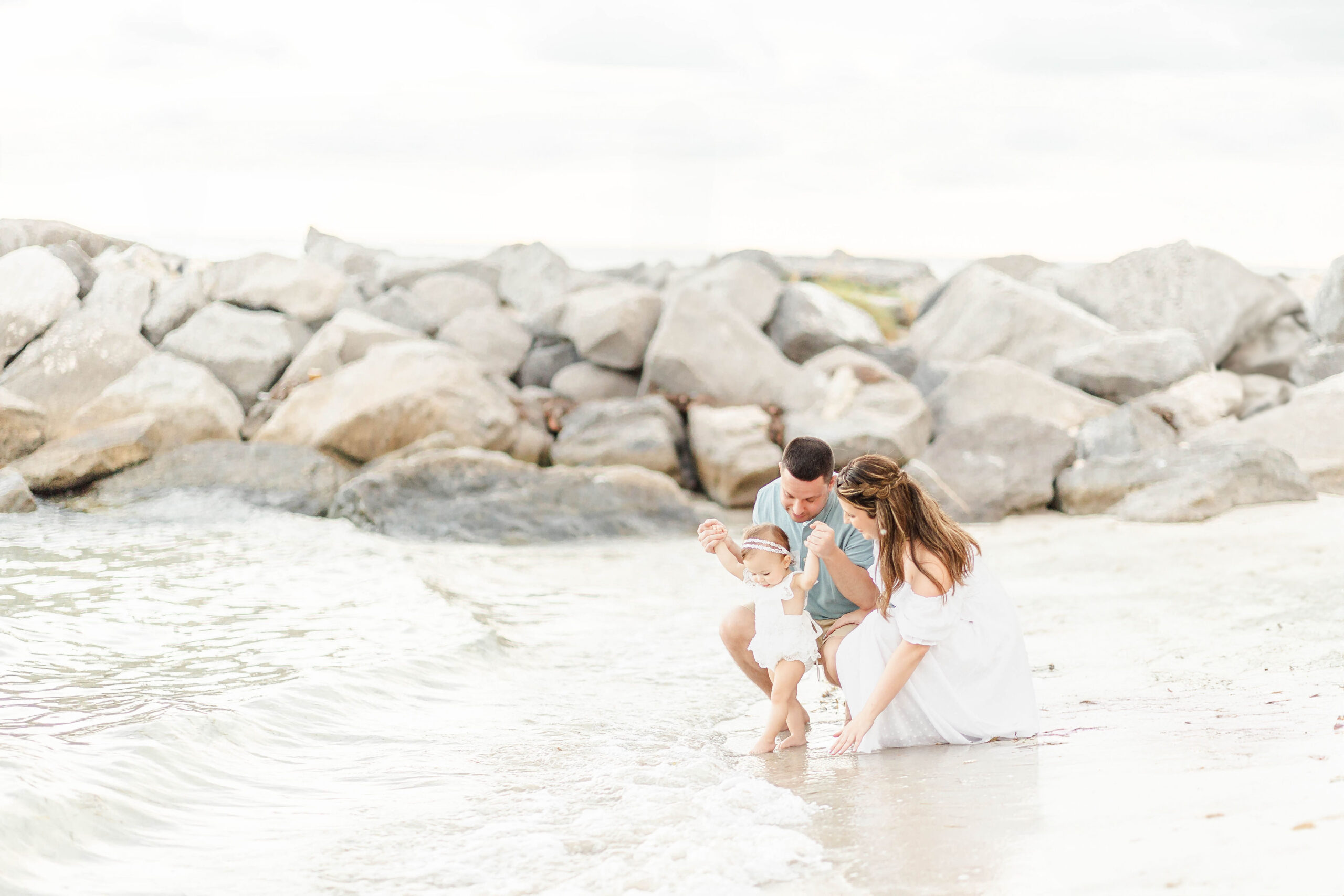 Mom and dad help their infant walk in the beach water in a white dress before visiting splash pads in Fort Lauderdale