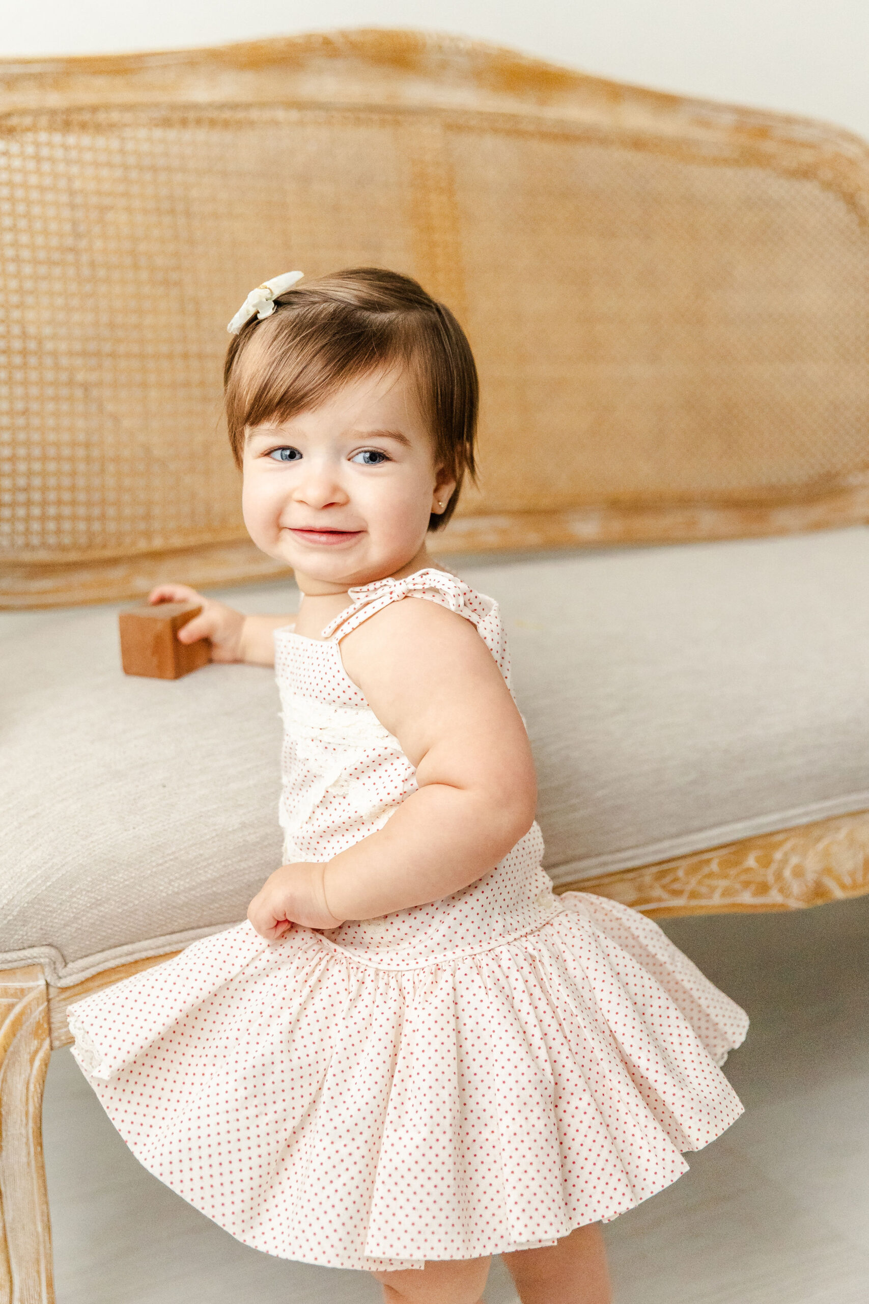 A happy baby girl in a white dress smiles over her shoulder while playing with a wooden block on a bench in a studio after some toddler activities in fort lauderdale