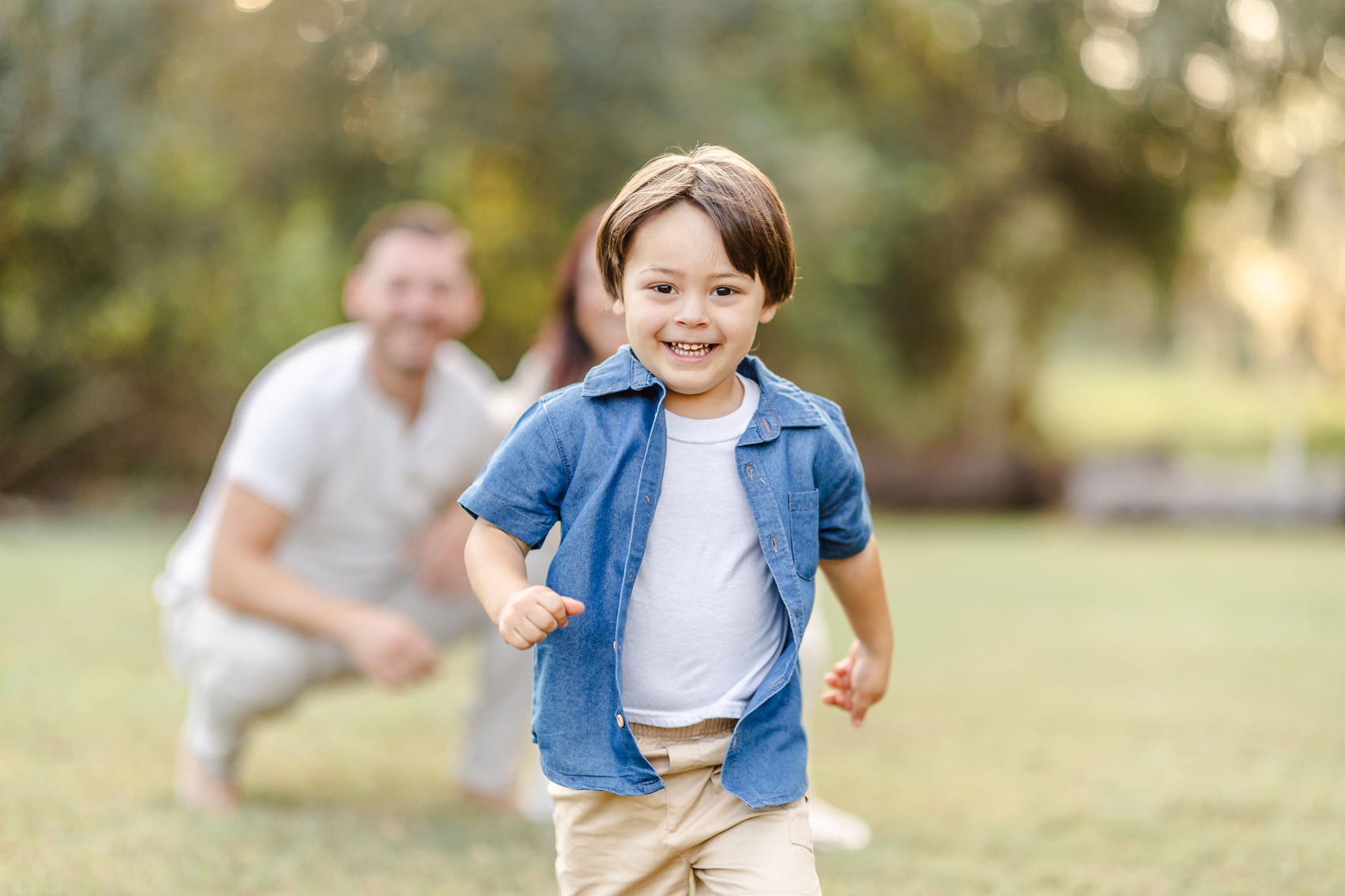 A giggling toddler boy in a denim shirt and khaki shorts runs in a park away from mom and dad