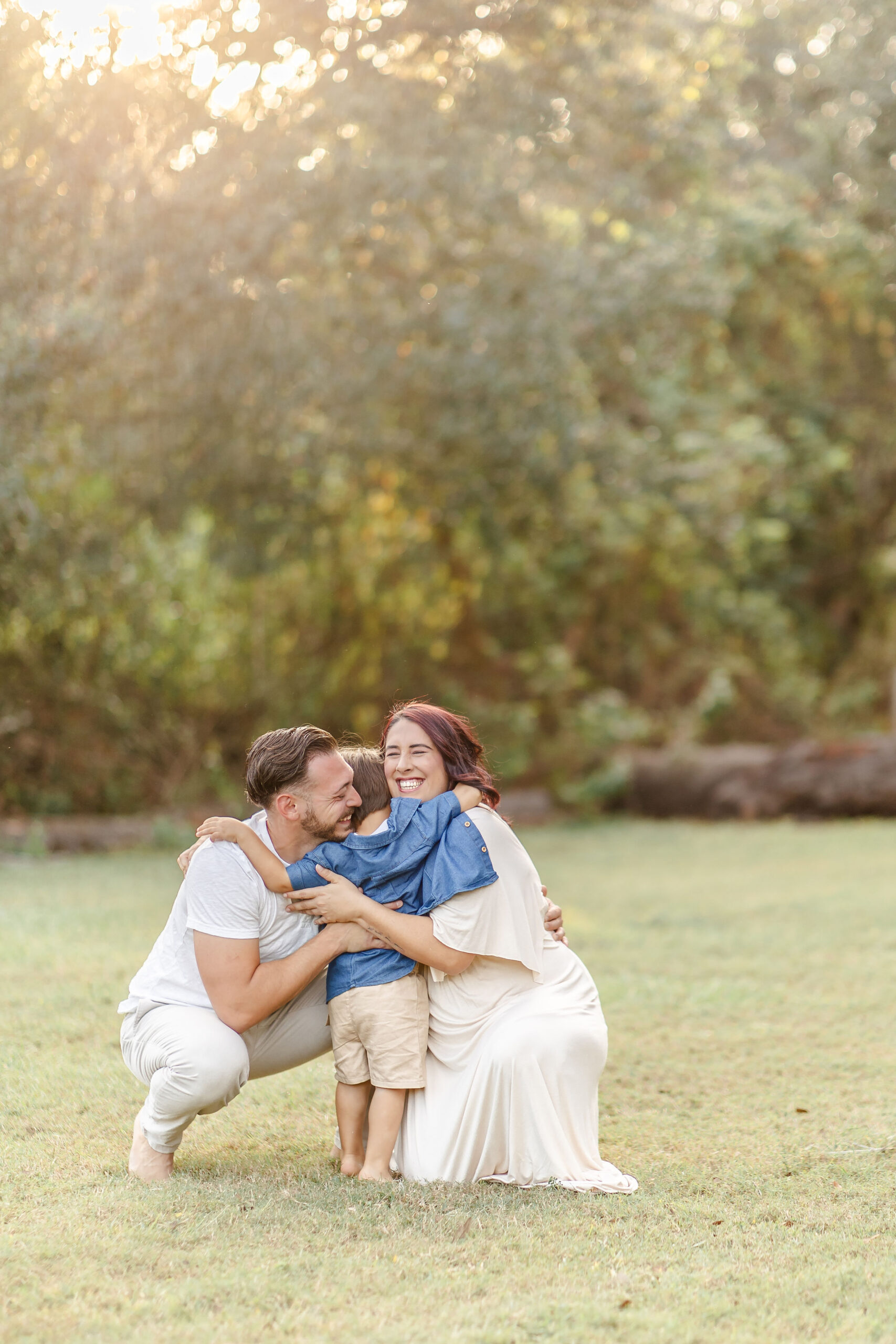 Happy mom and dad bend down for a big hug from their toddler son in a denim shirt in a park at sunset while staying at kid friendly hotels in fort lauderdale