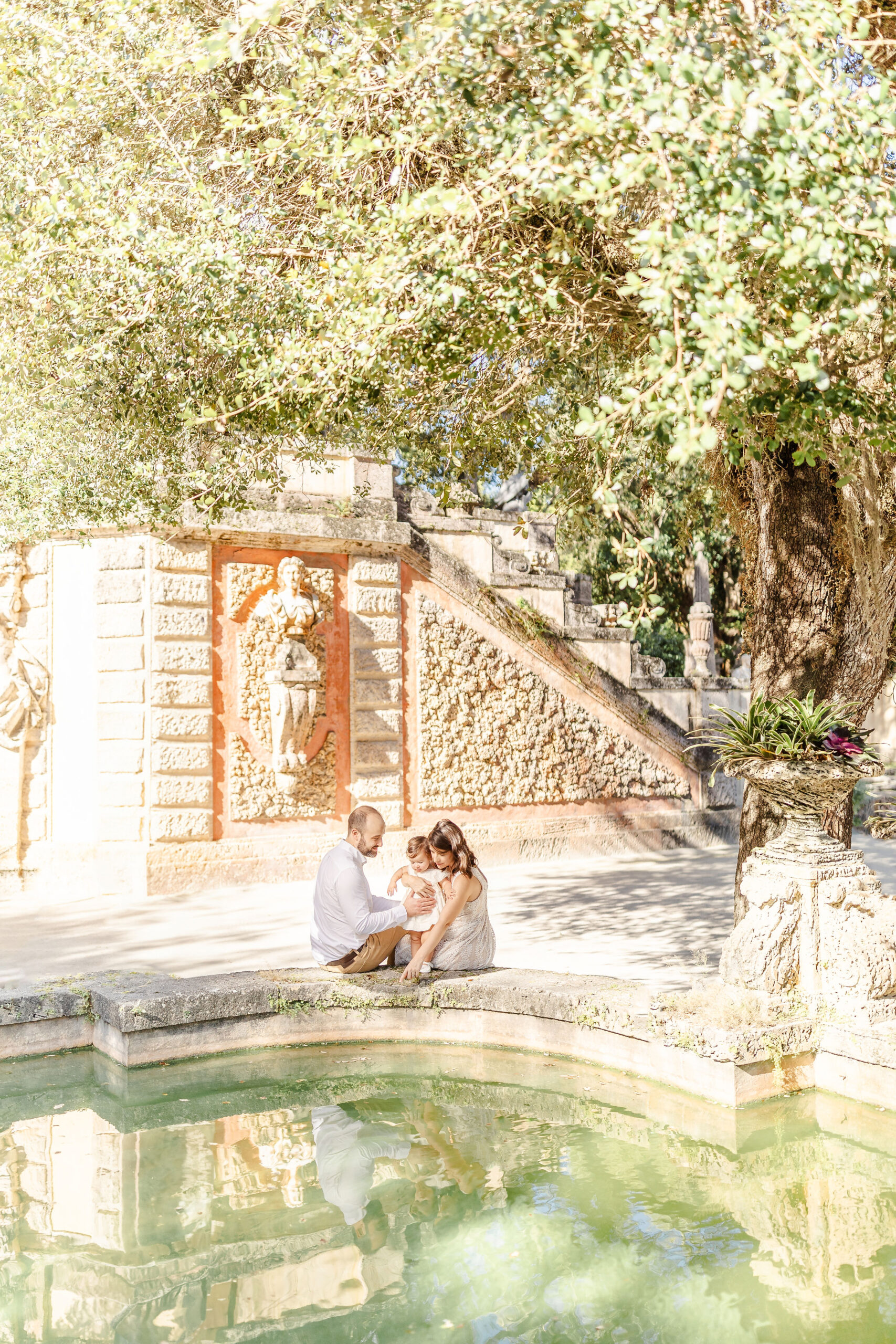 A mom and dad sit on the edge of a garden pond showing their toddler daughter the water