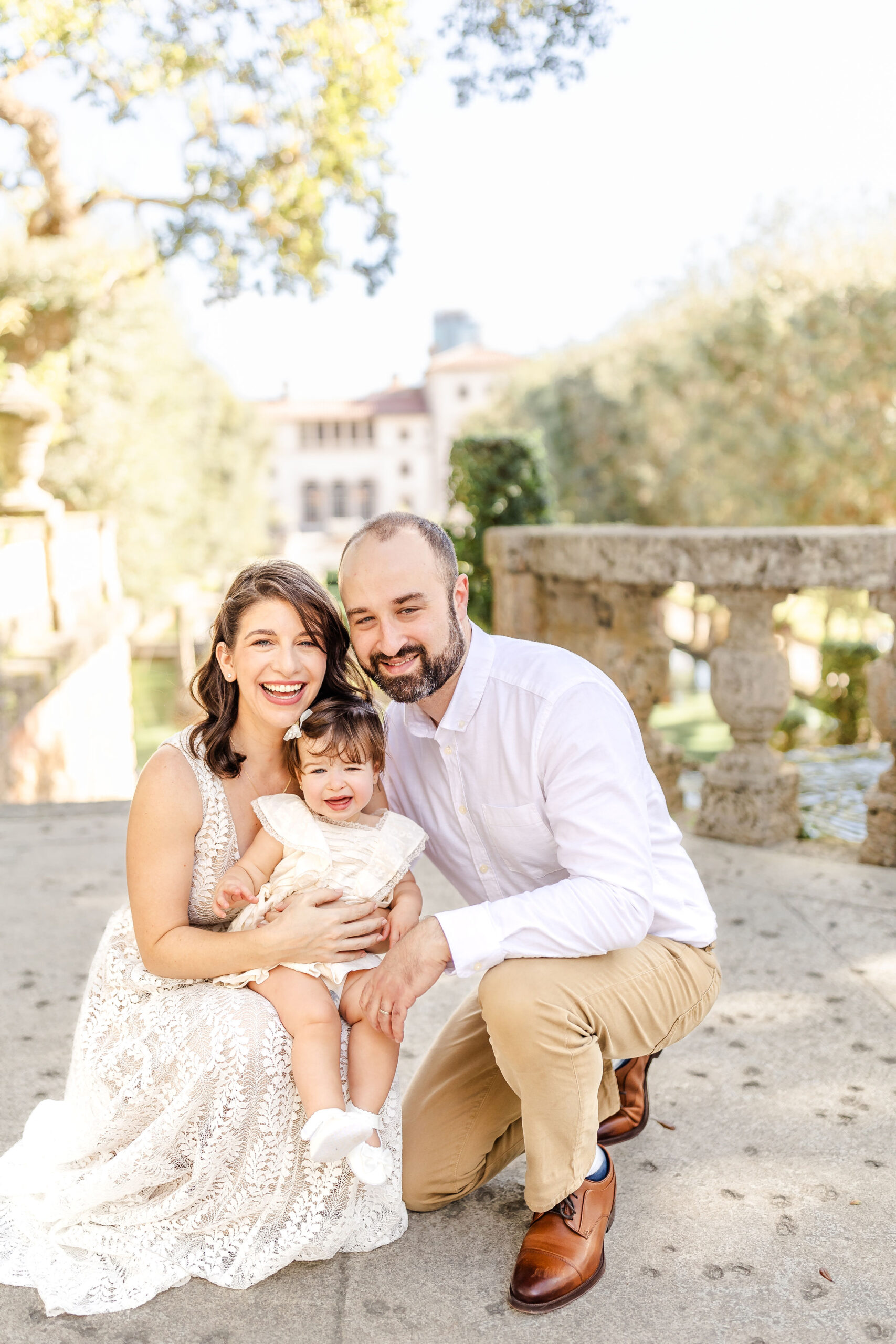Happy mom and dad kneel with their toddler daughter on mom's knee in a park at sunset before visiting kid friendly restaurants in fort lauderdale