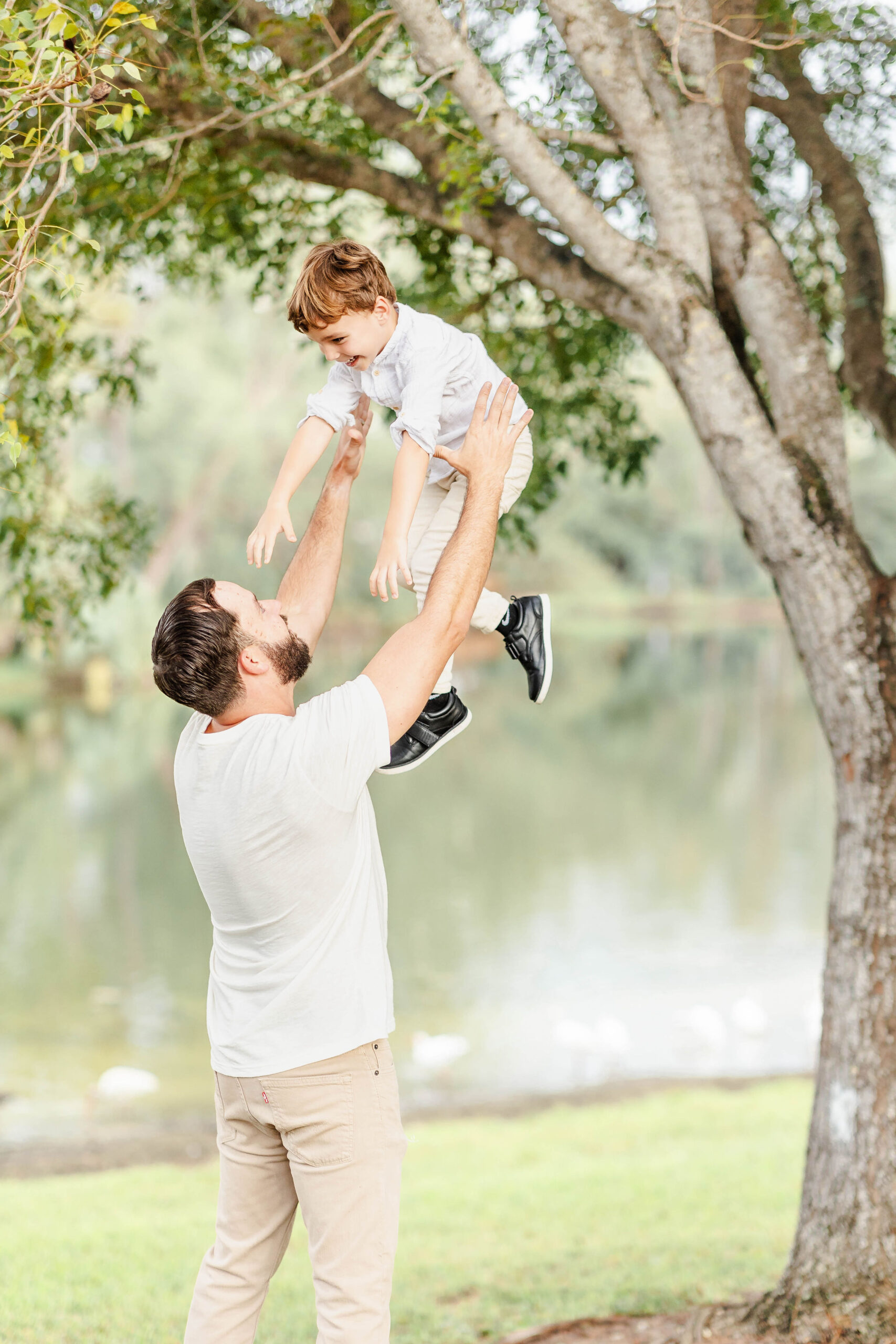 A dad in a white shirt tosses his young son in the air under a tree in a park