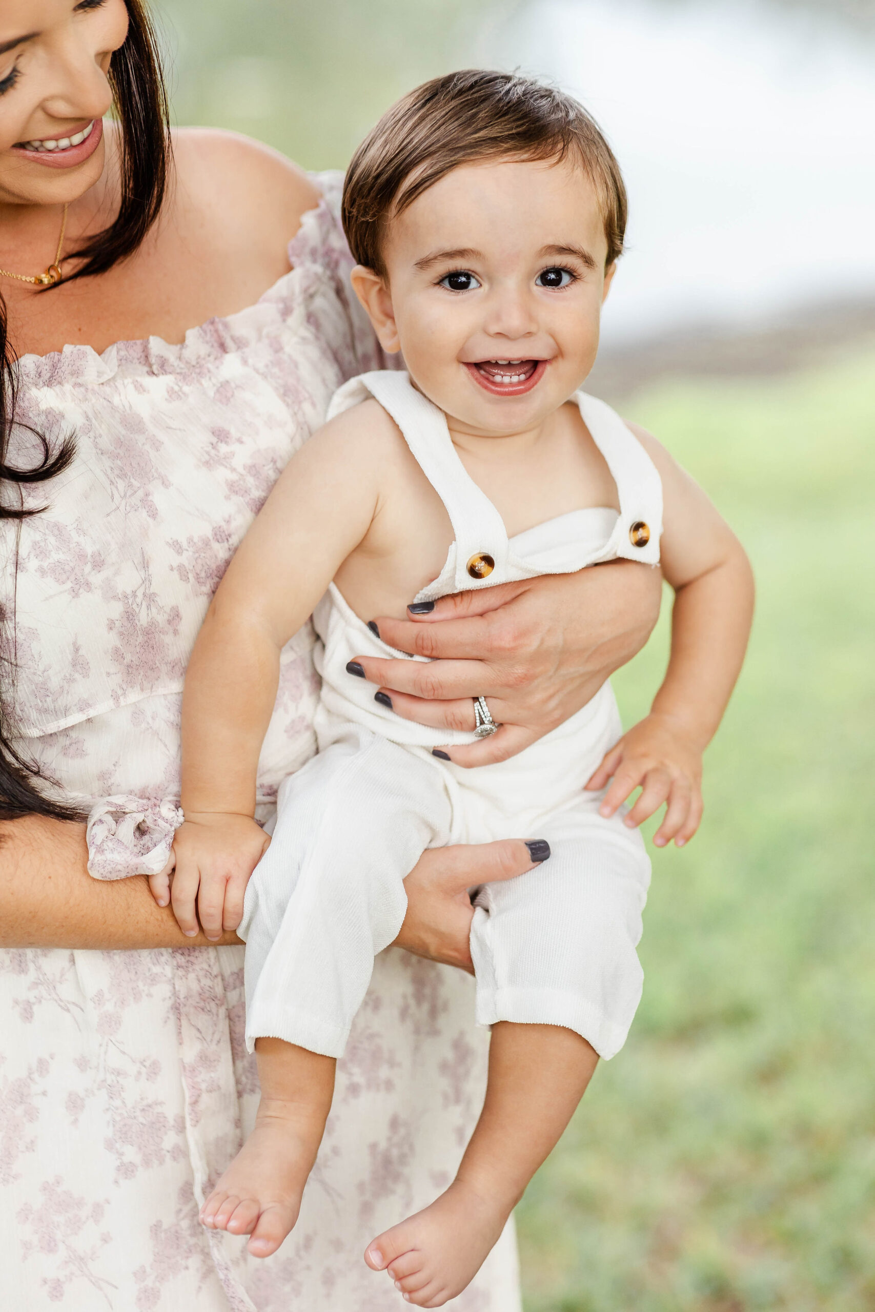 A giggling infant in white suspenders is held by mom in a pink floral print dress in a park before visiting playgrounds in fort lauderdale