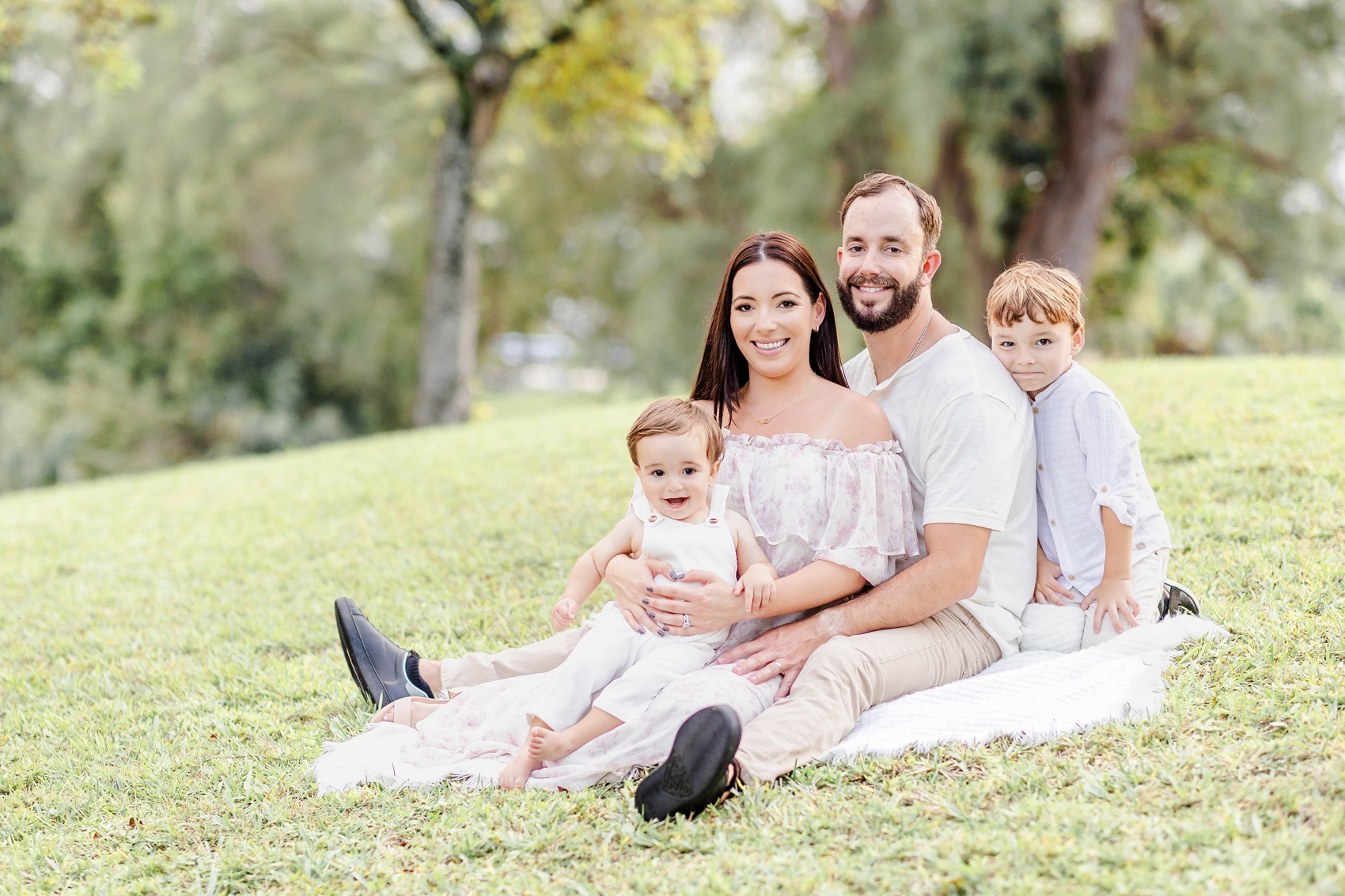Happy mom and dad sit on a picnic blanket in a park with their two happy toddlers before visiting playgrounds in fort lauderdale