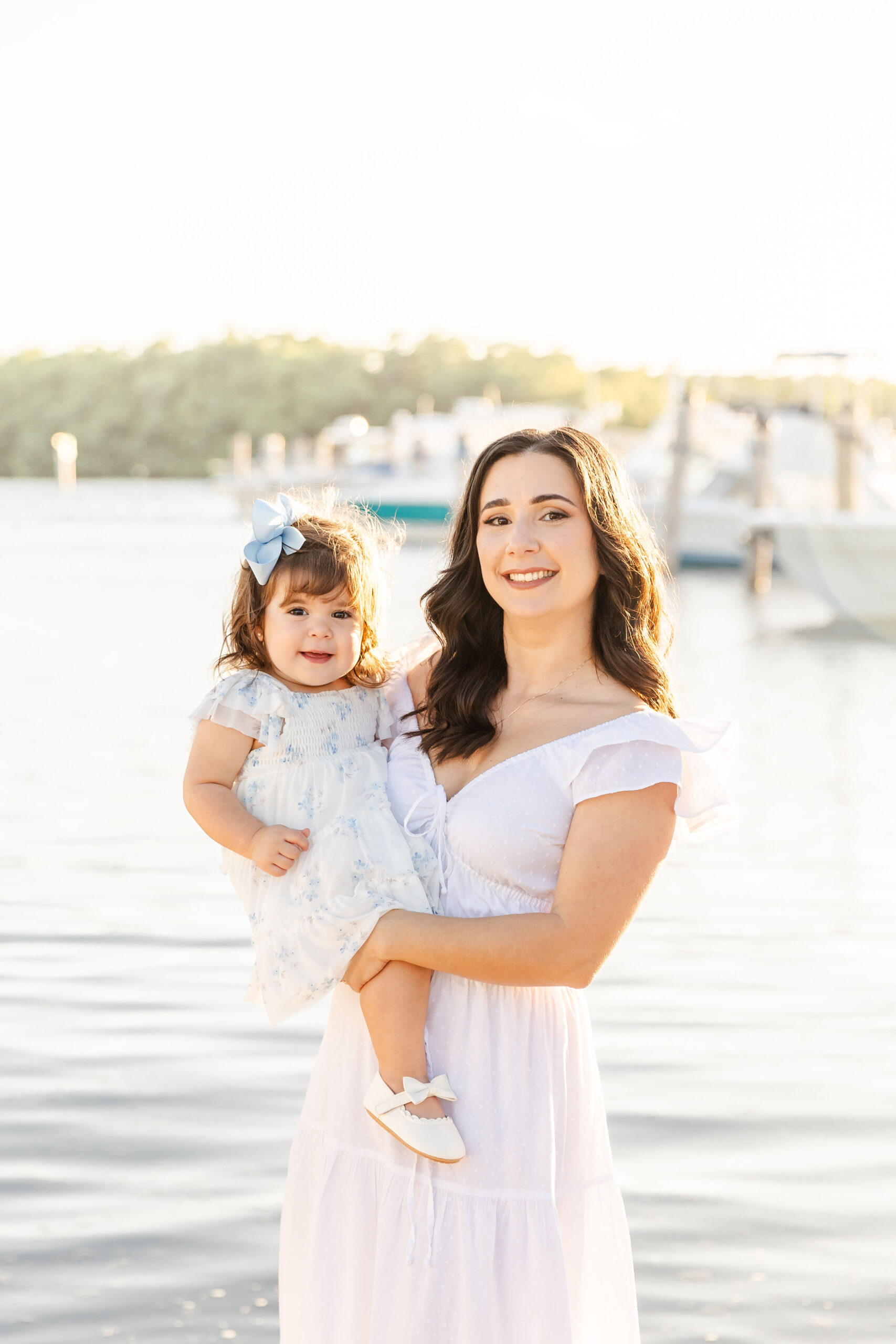 A happy mother in a white dress stands in a marina at sunset holding her smiling toddler girl in a floral print dress