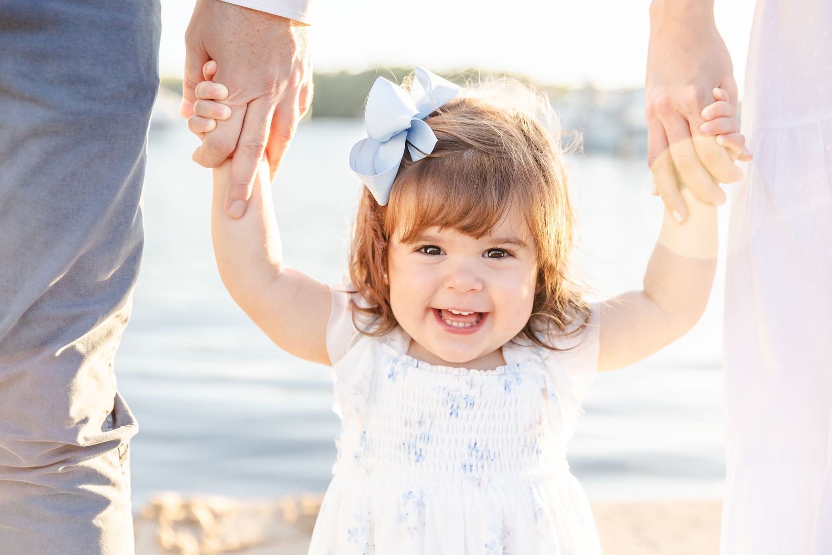 A happy toddler girl with a bow in her hair giggles while walking by the water holding mom and dad's hands in a white dress before visiting water parks near fort lauderdale